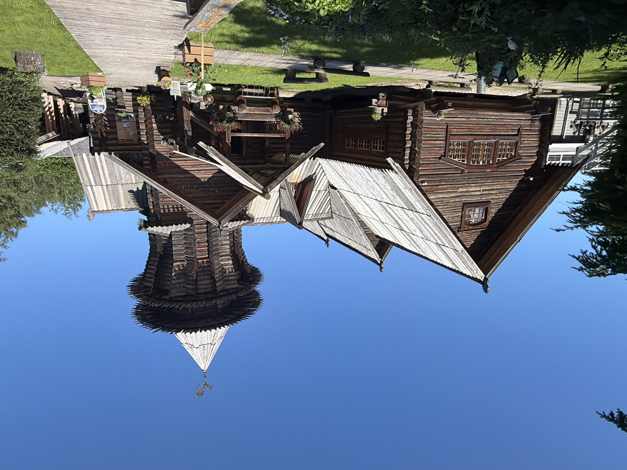 The image depicts a traditional wooden architectural complex under a clear blue sky. The main structure is a large, multi-gabled log building with a prominent, multi-tiered wooden tower topped with a weather vane shaped like a rooster. Several smaller wooden structures are attached or adjacent to the main building, all featuring weathered wooden roofs and log construction. A wooden walkway leads towards the entrance of one of the buildings. The grounds are covered in green grass and feature several wooden benches and planters with flowers. Trees and foliage are visible in the background and to the sides of the structures. The scene is illuminated by bright sunlight, casting shadows on the ground and the buildings.