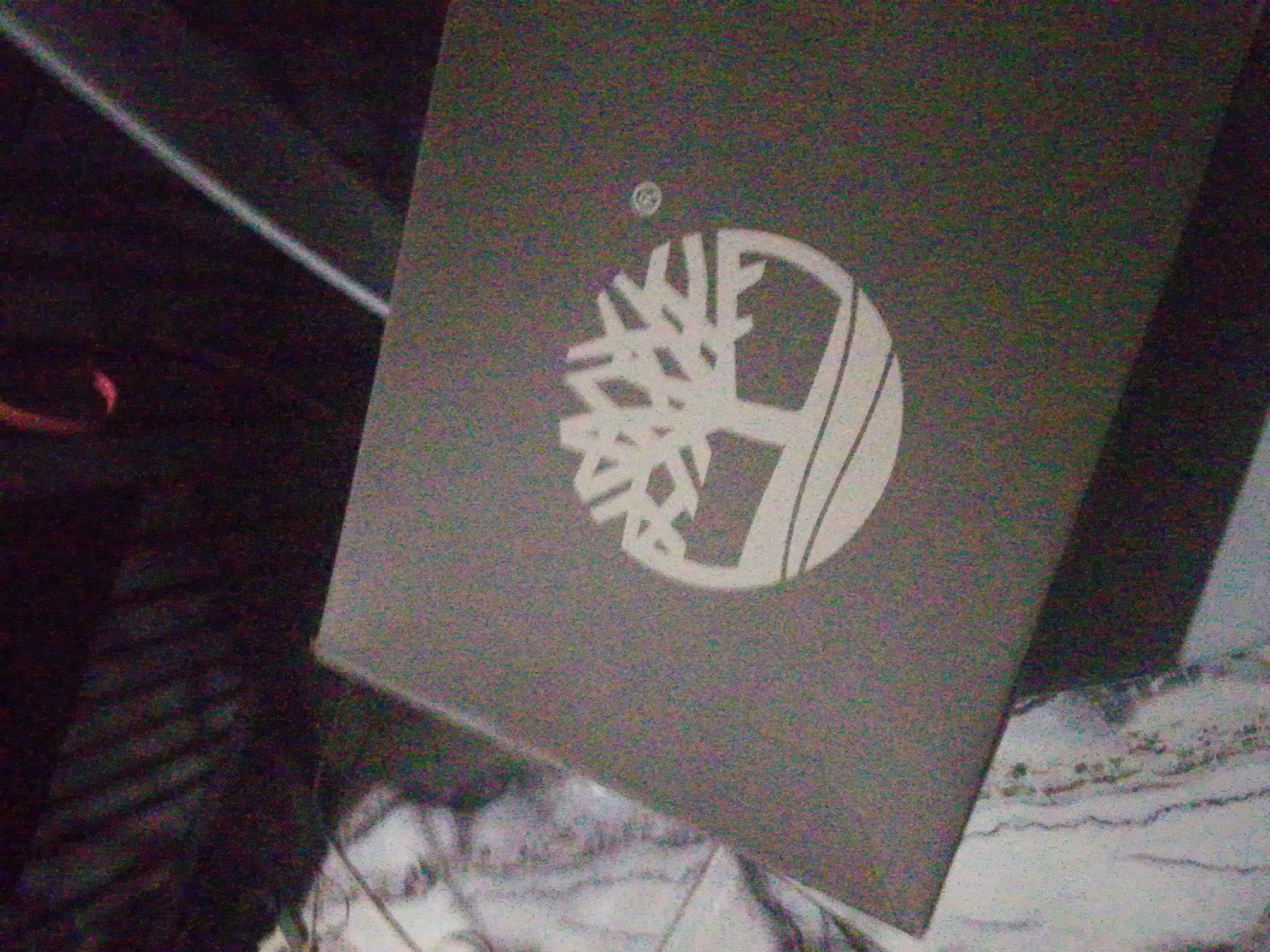 A dark grey rectangular object, likely a package or sign, is angled across the frame, displaying a prominent white Timberland logo. The logo consists of a stylized tree within a circular outline, topped by a small registered trademark symbol. The object rests on a lighter, mottled surface with indistinct wavy patterns in the lower right background, possibly resembling marble or textured fabric. The upper left background is dark and blurry, with a diagonal light-colored line and a small, blurred reddish element visible. The overall image exhibits significant digital noise and grain, indicating low-light conditions. No individuals or specific actions are depicted. The scene is located in the city of Unknown, Unknown.