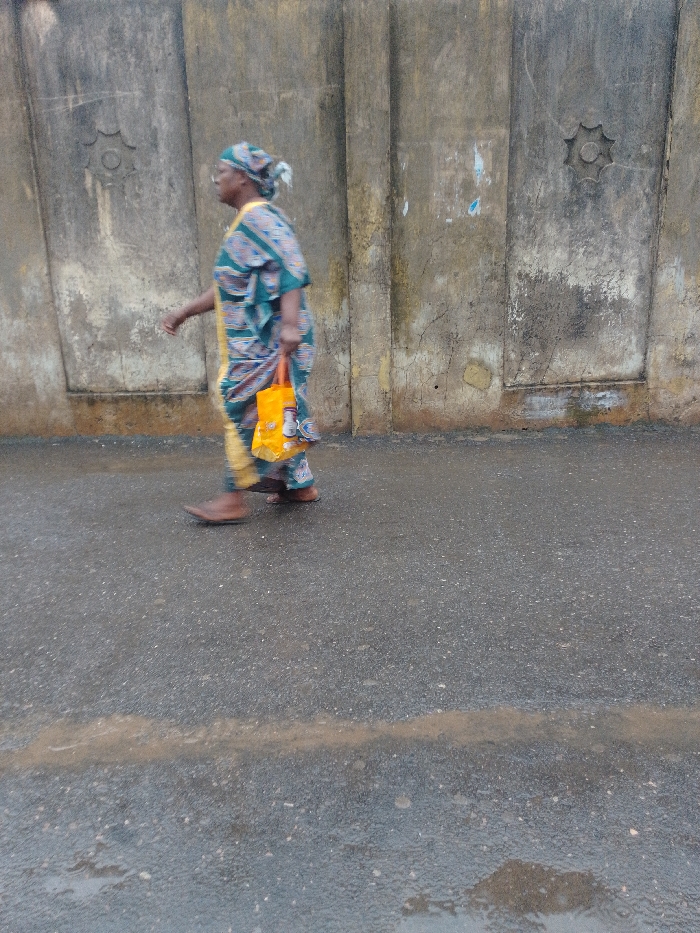 In Agani, Nigeria, a single adult female is observed walking from left to right on a damp, dark paved road. She is dressed in a patterned teal, blue, and yellow traditional garment, including a matching headwrap. Her left arm is slightly bent, and she carries an orange fabric bag in her right hand. Her footwear consists of brown shoes or sandals. The background features a weathered concrete or stucco wall with vertical indentations and two star-shaped decorative medallions. The road surface exhibits some light brown streaks or deposits. The lighting is diffused.