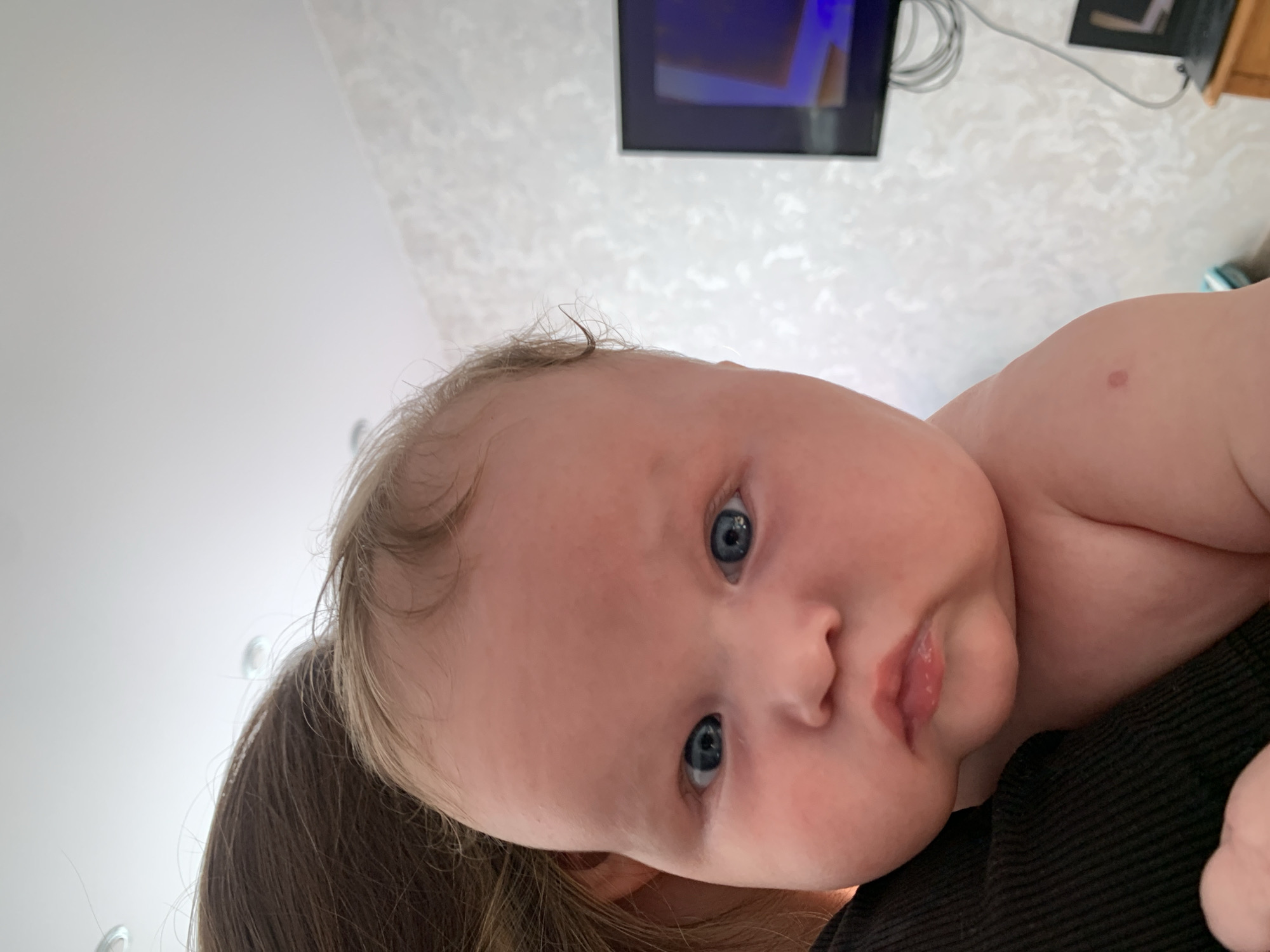This is a close-up, portrait-style photograph of a baby held by an unseen person. The baby is positioned to the right of the frame, with their face turned towards the camera. The baby has fair skin, light hair, and striking blue eyes. Their expression is curious and alert. A small, reddish birthmark is visible on the baby's upper arm or shoulder.
The background of the image is blurred but appears to be an indoor setting. A textured, light-colored wallpaper is visible on the wall behind the baby. In the upper portion of the image, a television screen displaying a brightly colored image and some electronic equipment, including cables and a wooden shelf, are partially visible. The overall lighting suggests daytime. The scene evokes a sense of intimacy and a personal moment between the baby and the person holding them. There is no visible text in the image.