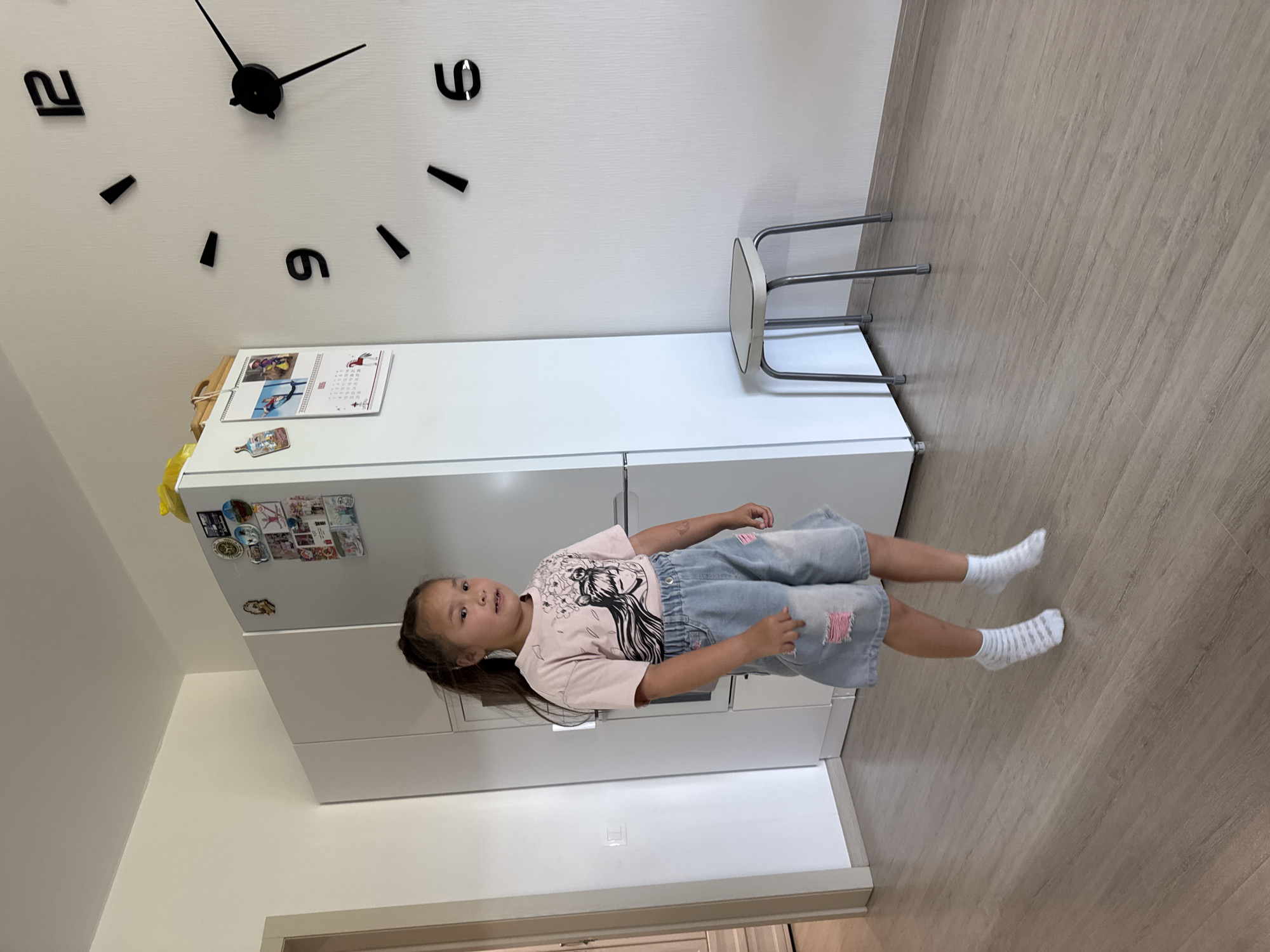 A young East Asian girl with dark hair in a ponytail, wearing a pink t-shirt and denim shorts, stands in what appears to be a kitchen or living area. The room has light-colored walls and wood-effect flooring. To the left of the girl is a tall, white refrigerator with magnets and pictures on its surface. To the right, a large modern clock with black numbers and hands is mounted on the wall, with a small stool positioned beneath it. The girl is looking towards the camera and her mouth is slightly open. She is wearing white socks with horizontal stripes. The overall lighting suggests it is daytime. The location is identified as Anapskaya, Russia.