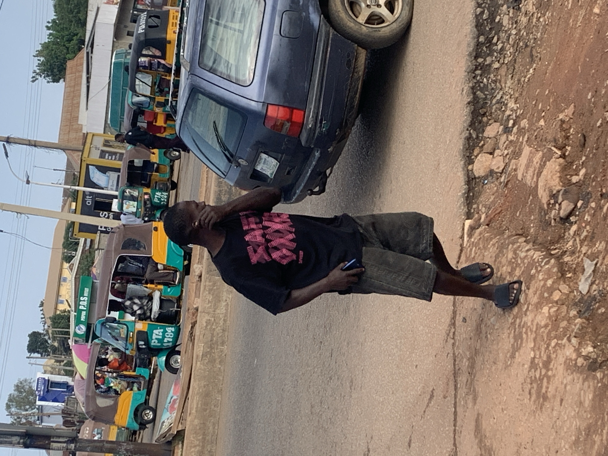In Gura Top, Nigeria, a man wearing a black t-shirt with pink lettering and gray shorts walks on a dirt road next to a busy street. He is holding a mobile phone in his left hand and has an earpiece in his right ear. The street is lined with various modes of transportation, including several yellow and green auto-rickshaws, commonly known as "Kekes." Some of these Kekes are filled with passengers, and one has a pink and green umbrella on top. A dark blue station wagon is parked on the right side of the street, partially obstructing the view of the road behind it. In the background, buildings and trees suggest a semi-urban environment. The sky is overcast.