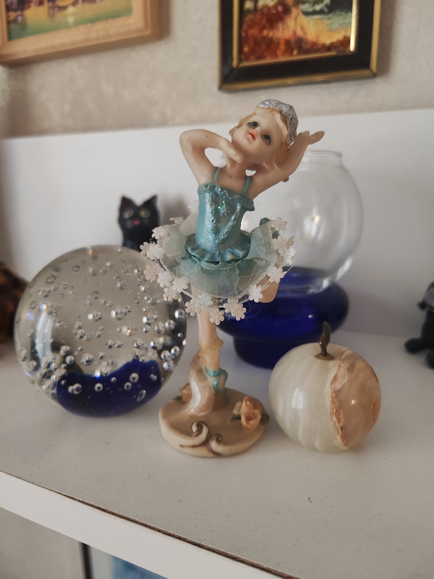 This image captures a close-up view of several decorative objects arranged on a white shelf. In the foreground, a small figurine of a ballerina in a blue tutu stands prominently, her arms gracefully extended. To the left of the ballerina is a clear glass orb filled with small bubbles and a dark blue object at its base. Behind the orb, a small, dark figurine of a cat with yellow eyes is visible. To the right of the ballerina, a smooth, white and tan polished stone in the shape of an apple sits on the shelf. In the background, a transparent glass bowl and a dark blue vase are partially visible. Above the objects, framed artwork can be seen hanging on a textured wall. This scene appears to be an indoor display of collectibles.