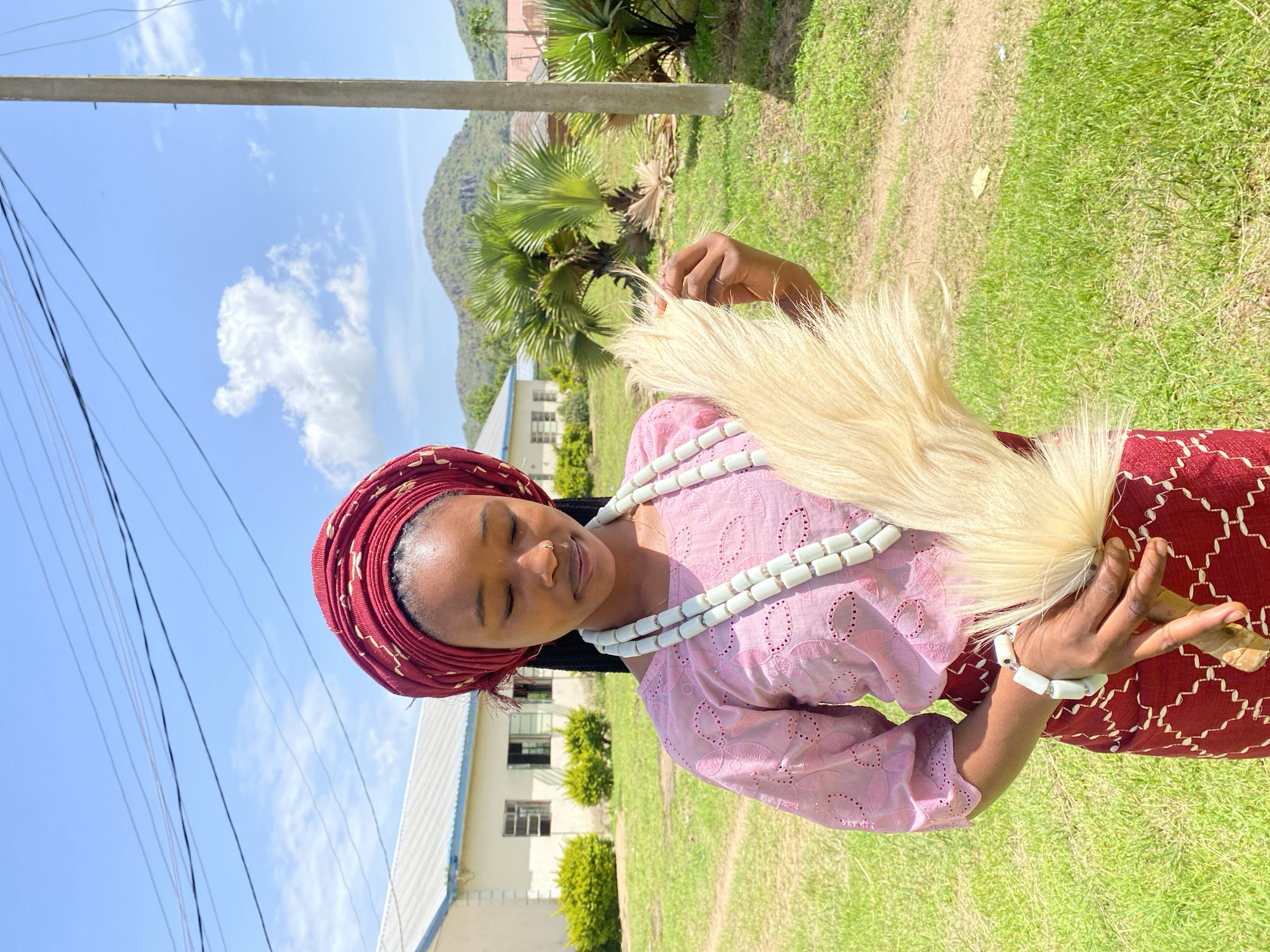 A young woman is photographed outdoors, wearing traditional attire. She has a red headwrap adorned with a gold pattern and a pink lace top. Around her neck, she wears multiple strands of white beads, and a bracelet of the same beads is on her wrist. In her hands, she holds a whisk-like object made of blonde, feathery material, with a wooden handle. Her eyes are closed, and she has a serene expression.
The setting appears to be in Vom, Nigeria. The background shows a building with a white facade and windows, indicative of an institutional or residential structure. Lush green grass covers the ground, with some bushes visible. In the distance, a hill or mountain can be seen under a bright blue sky with scattered white clouds. A utility pole and power lines are present in the foreground, suggesting the presence of infrastructure. The lighting indicates it is daytime, likely late morning or early afternoon, with bright sunlight casting shadows. The overall atmosphere is peaceful and evokes a sense of cultural tradition. No visible text is present in the image.