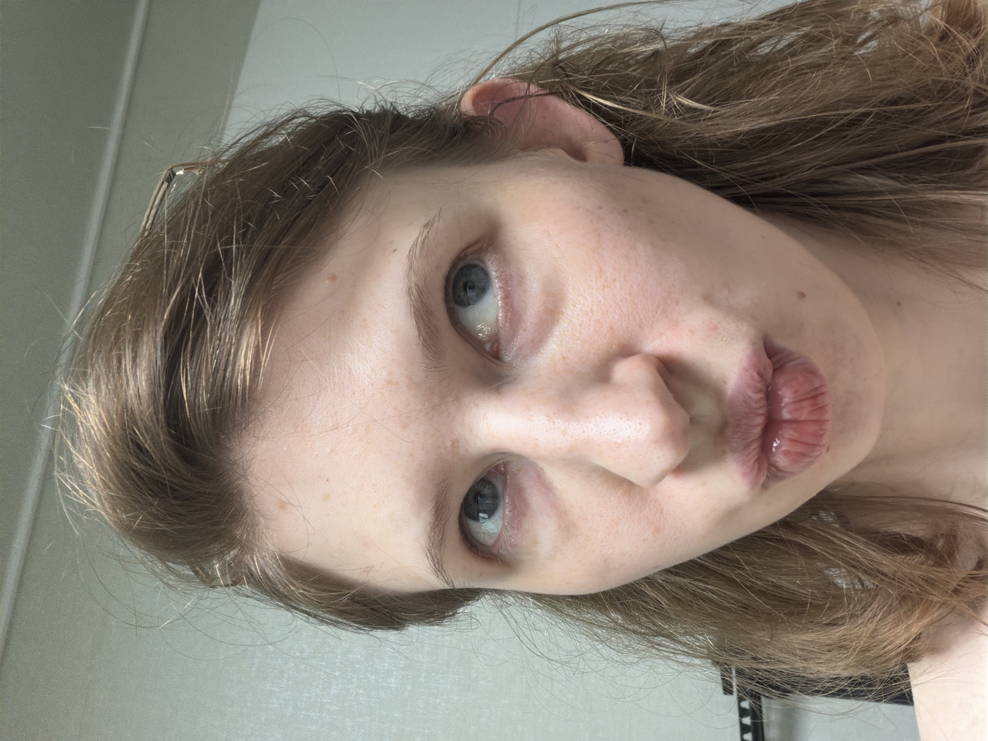 A close-up, head-and-shoulders shot focuses on a young Caucasian female with light brown hair, fair skin, and light blue eyes. She is looking upwards and slightly to her right, with her lips pursed as if to blow a kiss. A faint blush is visible on her cheeks, and a few freckles are scattered across her nose and forehead. Her hair is slightly tousled, with a few strands falling forward. She is wearing a clear, thin-rimmed pair of glasses perched on her head, partially obscured by her hair. The background is a plain, textured light-colored wall, with a hint of a dark object, possibly a shelf or frame, visible on the far left. The lighting is soft and even, casting no harsh shadows. The image appears to have been taken indoors.