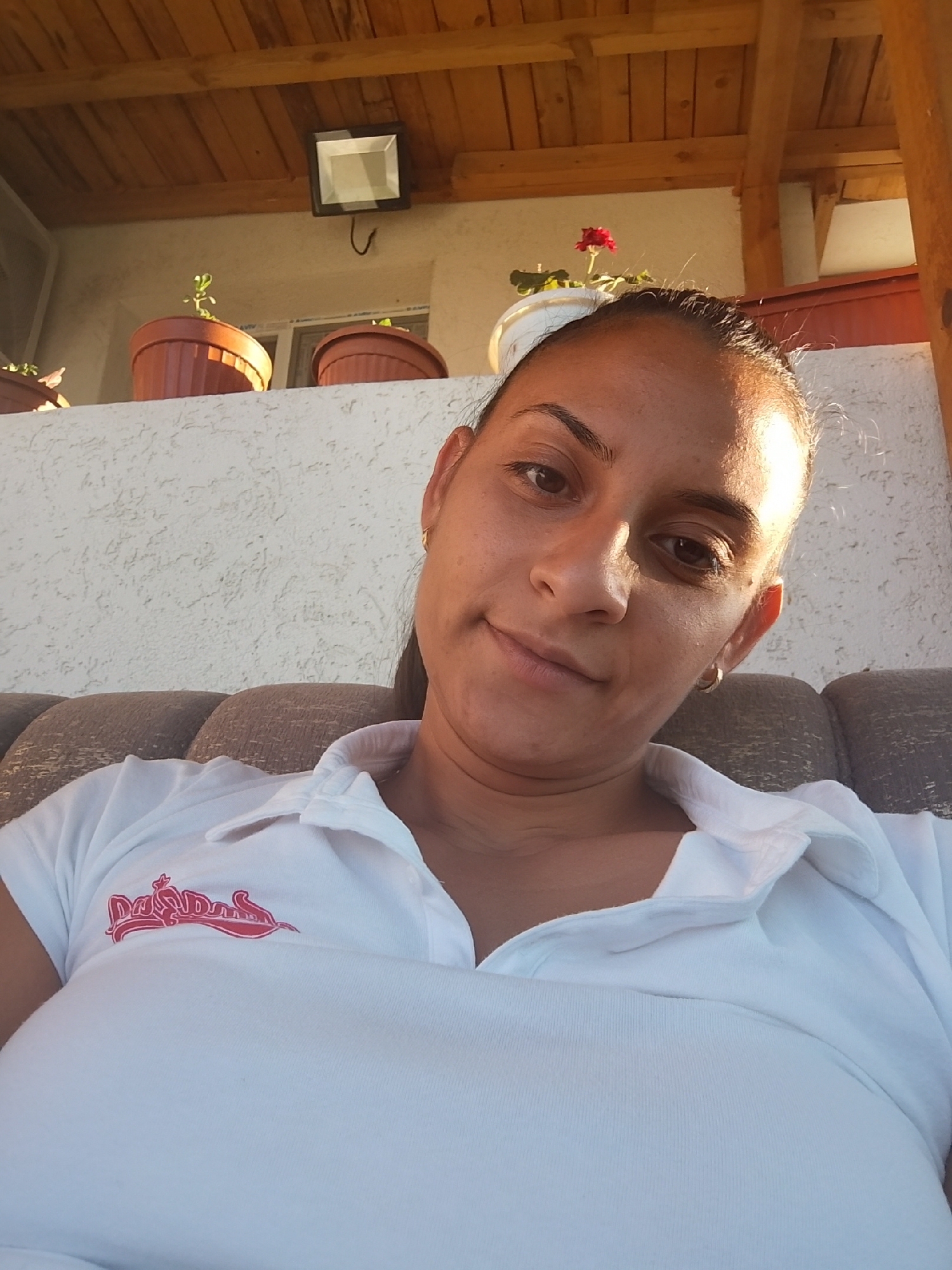 A medium shot selfie shows a woman with brown hair and brown eyes wearing a white polo shirt with a red logo on the left chest. She has a gold hoop earring in her left ear. The woman is outdoors, seated on a brown cushioned sofa. Behind her, a white textured wall is visible, and above that, a wooden-beamed roof. A black floodlight is mounted on the underside of the roof. Two potted plants are placed on a ledge against the wall. One pot is brown and contains a small green plant. The other pot is white and has a red flower. The scene is consistent with a residential outdoor area in Zimnitsa, Bulgaria.