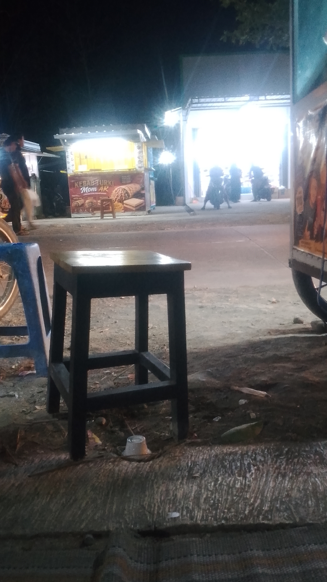 In Karanglangu, Indonesia, an outdoor scene unfolds at night. In the foreground, a dark wooden stool with a rectangular seat sits on the ground. To the left of the stool, a blue plastic chair is visible. Behind the stool, a food stall with a brightly lit display and signage that reads "KEBAB TURKI Mom AR" is set up. The stall features images of food items. Further in the background, the entrance to a building or another stall is illuminated by bright lights, with several people and motorcycles present. The ground is a mix of dirt and asphalt. The overall ambiance suggests a street-side dining or vending area at night.