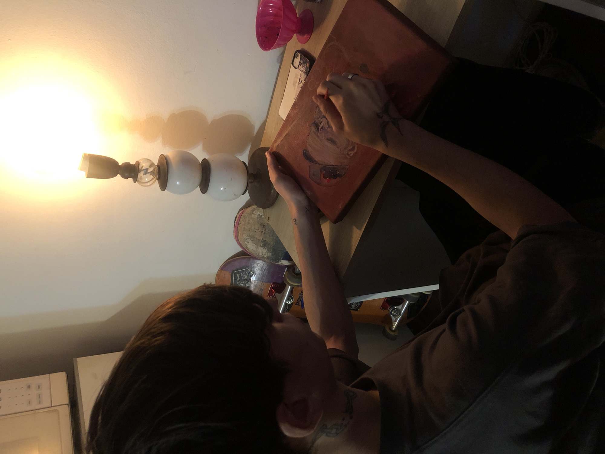 A person with dark hair and tattoos on their forearms is sitting at a desk and working on a painting. The painting, which is reddish-brown, shows a figure wearing a bowl-shaped hat. On the desk is a pink ice cream cup, a cell phone, and a lamp with a bright light. Under the desk are multiple skateboards. The location appears to be indoors in Buenos Aires, Argentina. The person is interacting with the painting, seemingly making adjustments or adding details.