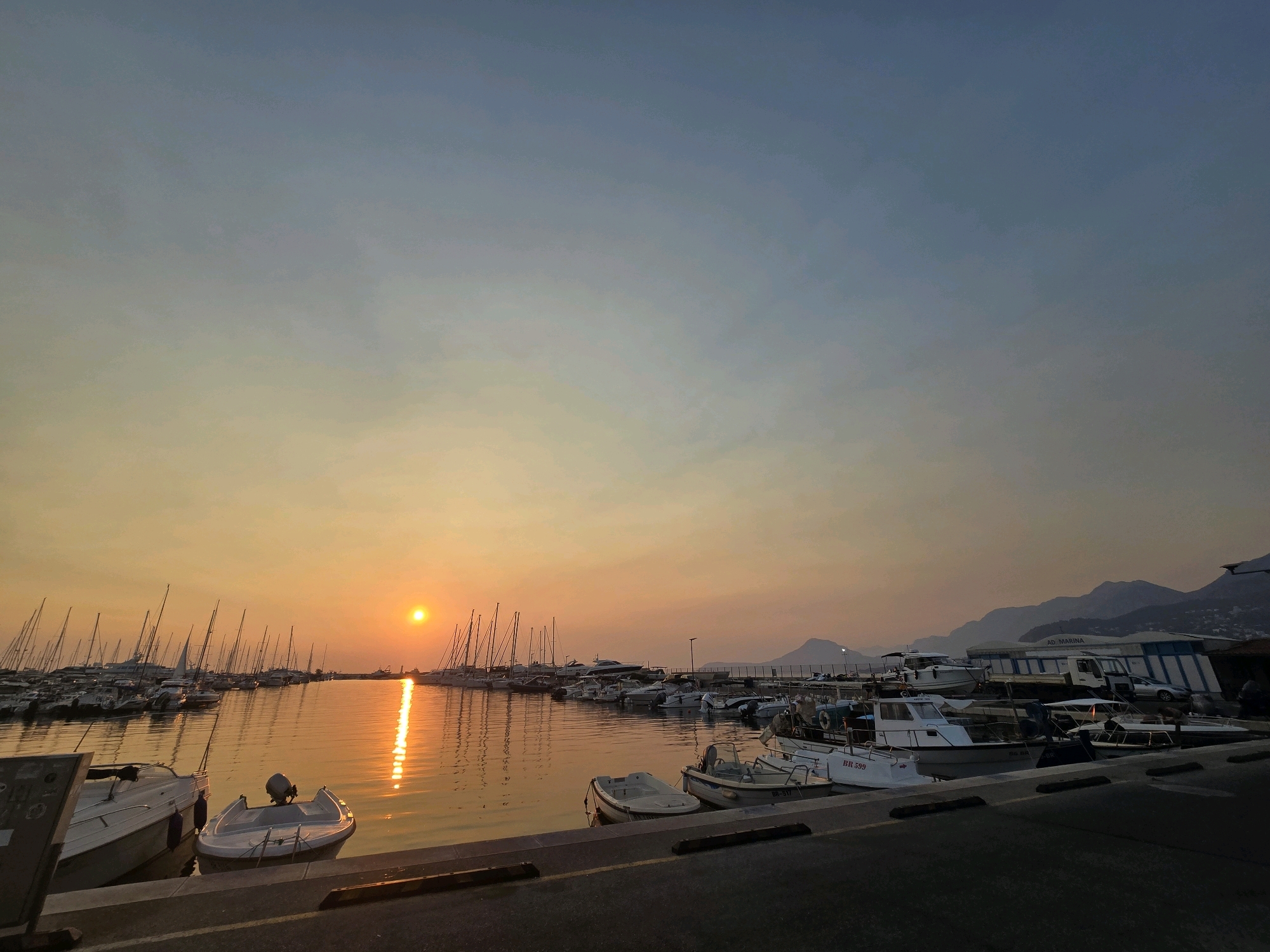An image captured at sunset in Bar, Montenegro, shows a tranquil marina scene. The sky transitions from a soft orange glow near the horizon to a dusky blue overhead, with the sun a bright orb reflecting a shimmering path across the calm water. Numerous sailboats and smaller motorboats are moored in neat rows, their masts creating a delicate silhouette against the vibrant sky. In the foreground, a paved walkway borders the water, with a few smaller boats tied up along the edge. Further back, buildings line the shore, some with visible signage, and mountainous terrain forms a hazy backdrop. The scene is serene and quiet, devoid of any visible people or activity.