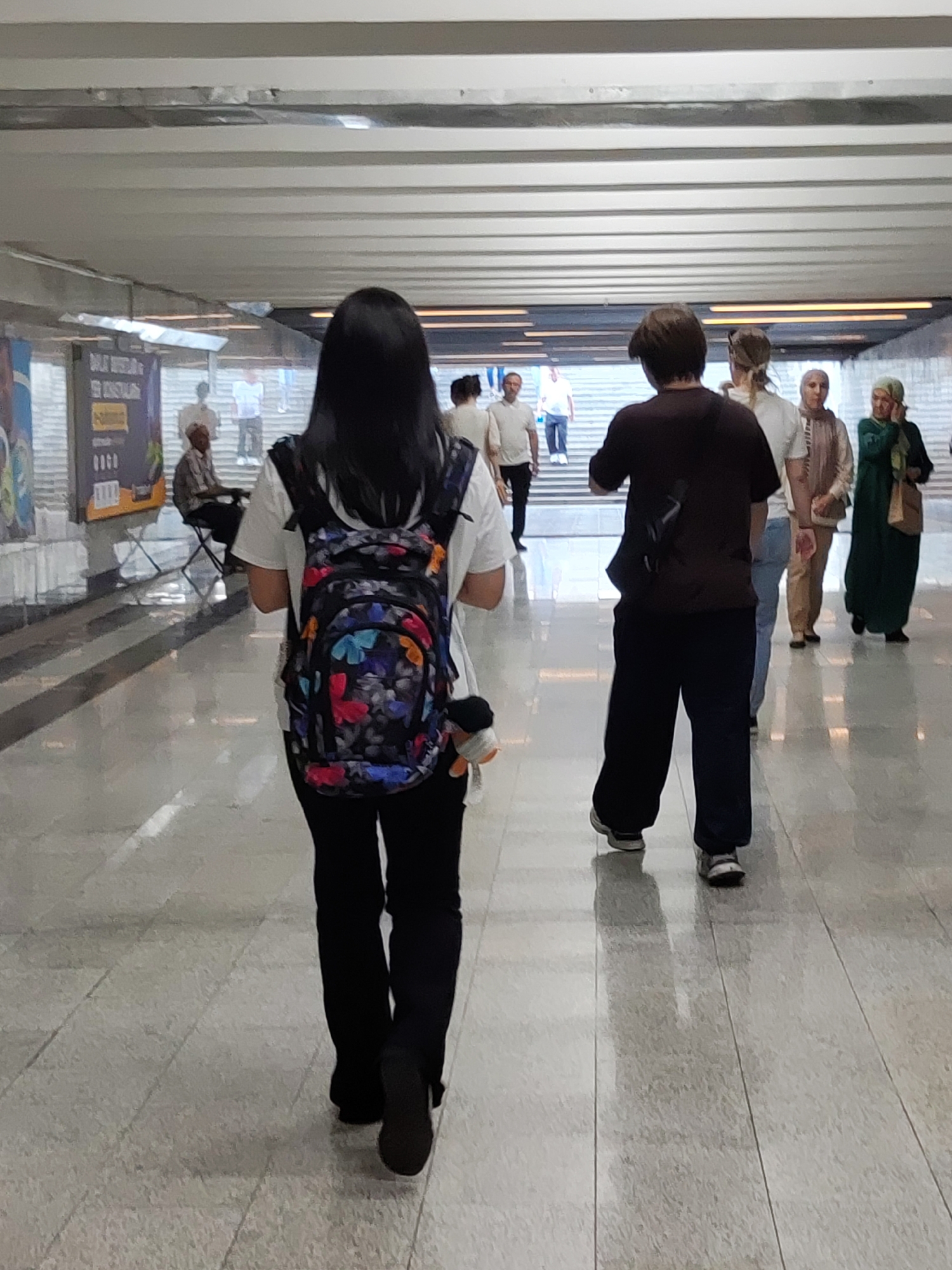 The image depicts a view from behind a young woman with long black hair wearing a white t-shirt, black pants, and a colorful backpack with a butterfly pattern. She is walking down a well-lit, modern pedestrian underpass. To her right, a man in a dark t-shirt and dark pants walks ahead of her. Further ahead, a group of people are visible, including two women wearing hijabs and another person. On the left side of the underpass, a man is seated at a table, and a poster is displayed on the wall. The underpass appears to be a busy thoroughfare, possibly part of a metro station or a public transport hub in Tashkent, Uzbekistan, characterized by its clean, tiled flooring and overhead lighting system.