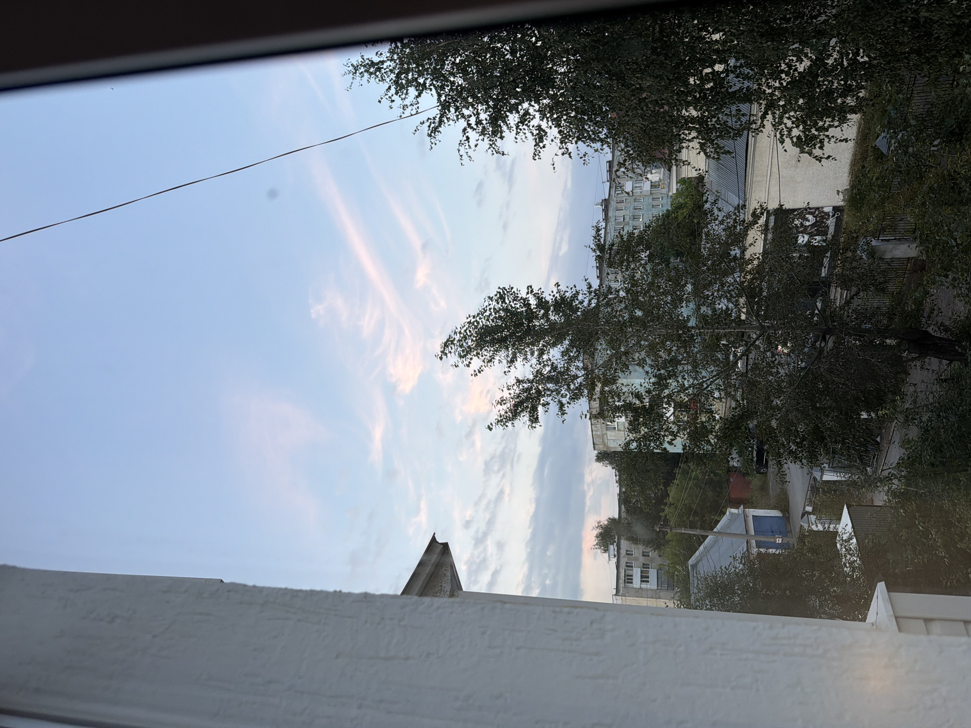 The image is a view from a window in Lysva, Russia, looking out over a residential area. The sky is a pale blue with wispy pink clouds, suggesting either dawn or dusk. In the foreground, a white textured wall and a window frame are visible on the left, partially obscuring the view. Beyond the immediate window, trees with dark green foliage dominate the middle ground, with some branches extending into the frame. Behind the trees, several apartment buildings are visible, their facades a mix of light blue and white with visible windows. A smaller building with a blue door and a gray roof is situated in the middle ground, possibly a garage or shed. A utility pole with wires is also present. The overall scene depicts a typical urban landscape in Russia, characterized by apartment blocks, trees, and utility infrastructure under a soft, atmospheric sky.