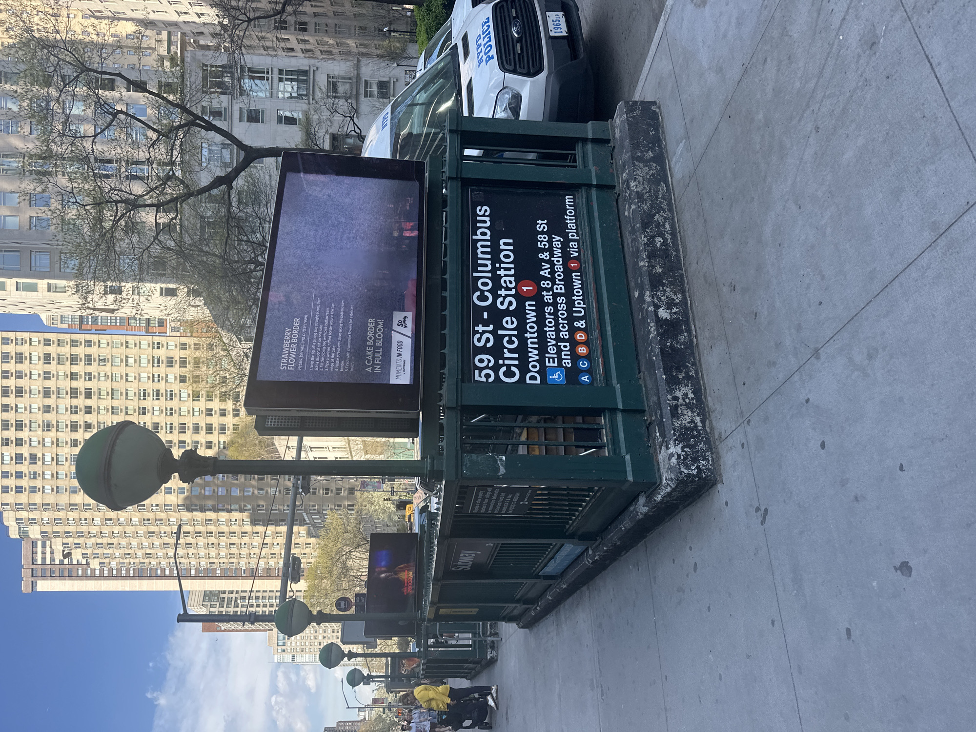 The image depicts a street scene in New York City, United States, featuring an entrance to the 59 St-Columbus Circle subway station. The subway entrance is marked with a green sign displaying the station's name and information about train lines (1, A, C, B, D) and elevator access. Adjacent to the station entrance is a digital advertising display.
In the background, there are tall buildings and trees lining the street. A New York Police Department (NYPD) vehicle is parked near the subway entrance. Pedestrians are visible on the sidewalk in front of the subway entrance. Street lamps are also seen in the scene.