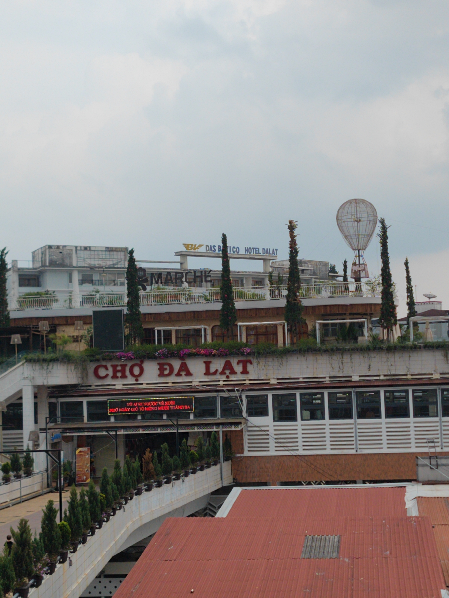 The image shows a multi-story building complex in Da Lat, Vietnam, identified by the large sign "CHỢ ĐÀ LẠT" (Da Lat Market) in red letters. Above the market, the buildings feature additional businesses, including "DAS BATICO HOTEL DALAT" and "MARCHE." A decorative hot air balloon structure is visible on the roof level. The sky is overcast. A pedestrian bridge, lined with potted plants, leads towards the market. Several different types of tall, green trees are also seen.