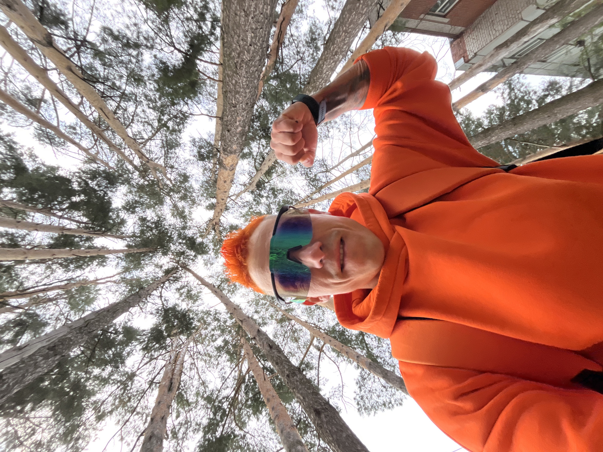 An outdoor, low-angle, wide shot shows a man with bright orange, spiky hair and neon green and blue mirrored sunglasses posing for a selfie. He is wearing an orange hooded sweatshirt with a backpack strap visible over his left shoulder. His right arm is flexed, showing his bicep. The man is surrounded by tall pine trees, their trunks extending upwards and their branches creating a canopy against a cloudy sky. A brick building is visible in the background on the right side of the frame. The image was taken in the city of Chaikovsky, Russia.