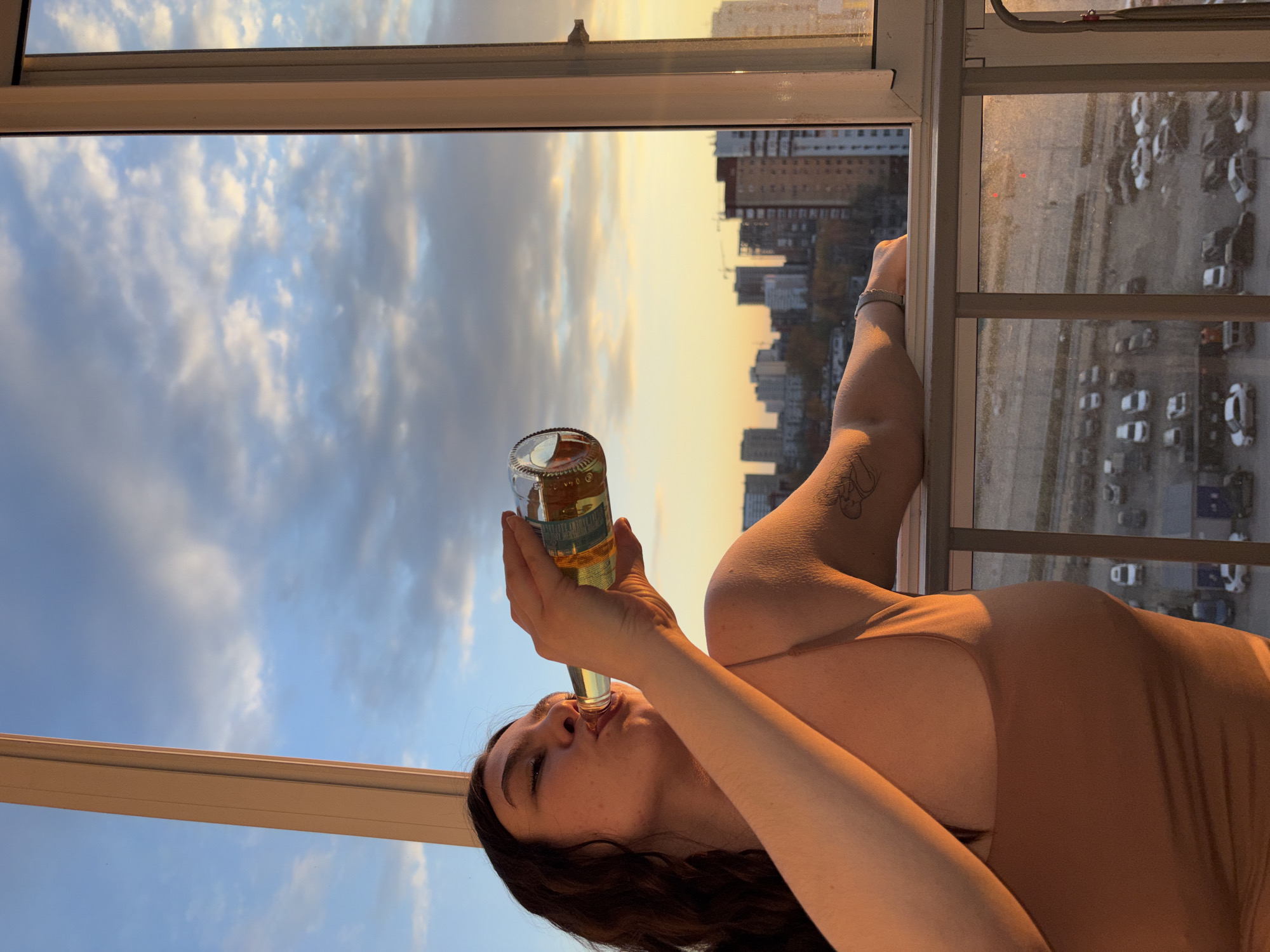 The image depicts a woman drinking from a clear glass bottle while leaning against a window frame, with an urban skyline visible outside at sunset. The original image is rotated 90 degrees clockwise, so this description assumes a corrected upright orientation.
The woman, with dark hair, is shown from the chest up on the right side of the frame. She wears a light-colored (possibly beige or nude) top. Her right arm, adorned with a tattoo on the upper arm and a bracelet on the wrist, holds the bottle to her mouth. Her eyes are closed or looking upward as she sips from the bottle, which contains a yellow or golden liquid.
The window frame, light-colored and prominent, acts as a boundary between the indoor setting where the woman is and the exterior urban landscape. Through the window, a sprawling city is visible under a vibrant sunset sky. The sky displays a blend of warm orange, yellow, and blue tones with scattered white and grey clouds, characteristic of dusk or golden hour. Numerous tall buildings create a dense skyline in the middle ground, while a large parking lot filled with many cars is visible below them. The warm, low-angle light of the setting sun illuminates the scene, casting a golden glow on the woman and parts of the cityscape. No discernible text is legible on the bottle's label. The overall scene suggests a moment of relaxation at a high vantage point overlooking the city of Perm, Russia.