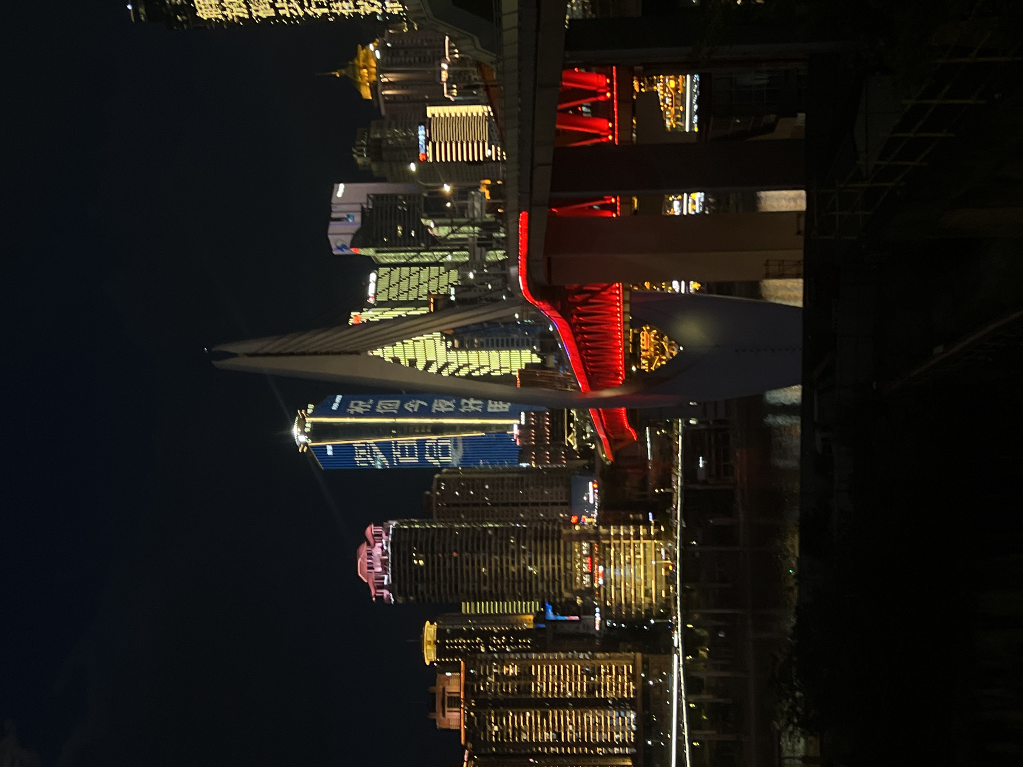 The image captures a nighttime cityscape of Jiangbeicheng Street, China, dominated by illuminated skyscrapers and a prominent bridge. The foreground features a dark, silhouetted landscape with some hints of vegetation and a section of a railing. A large, modern bridge with bright red lights spans across the frame, with its supporting pillars visible. Beyond the bridge, a dense collection of high-rise buildings lights up the night sky. One particularly tall, blue building in the middle ground displays vertical white lettering. To the left and right of this building are other skyscrapers with various architectural styles and illuminated windows. The scene is a vibrant display of urban nightlife, with the city's architecture and infrastructure illuminated against the dark night.