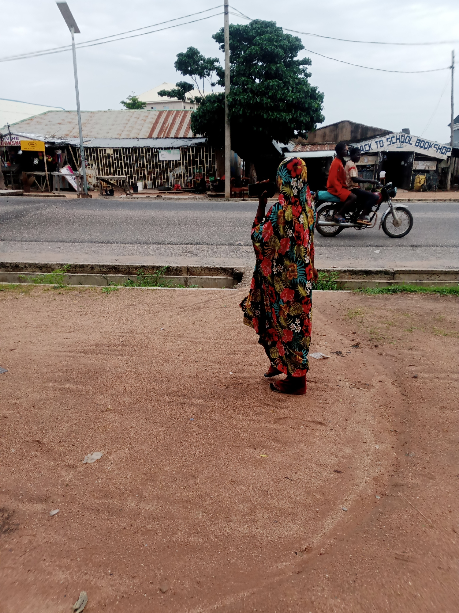 A woman wearing a colorful floral dress stands in the foreground, facing away from the camera. She is on a dirt path next to a paved road. Behind the road, several buildings line the street. One building has a corrugated metal roof and a sign that reads "DIVINE BOOKSHOP". Further down the street, another building has a sign that says "BACK TO SCHOOL BOOK SHOP". A motorcycle carrying two people is riding on the road. The sky is overcast.