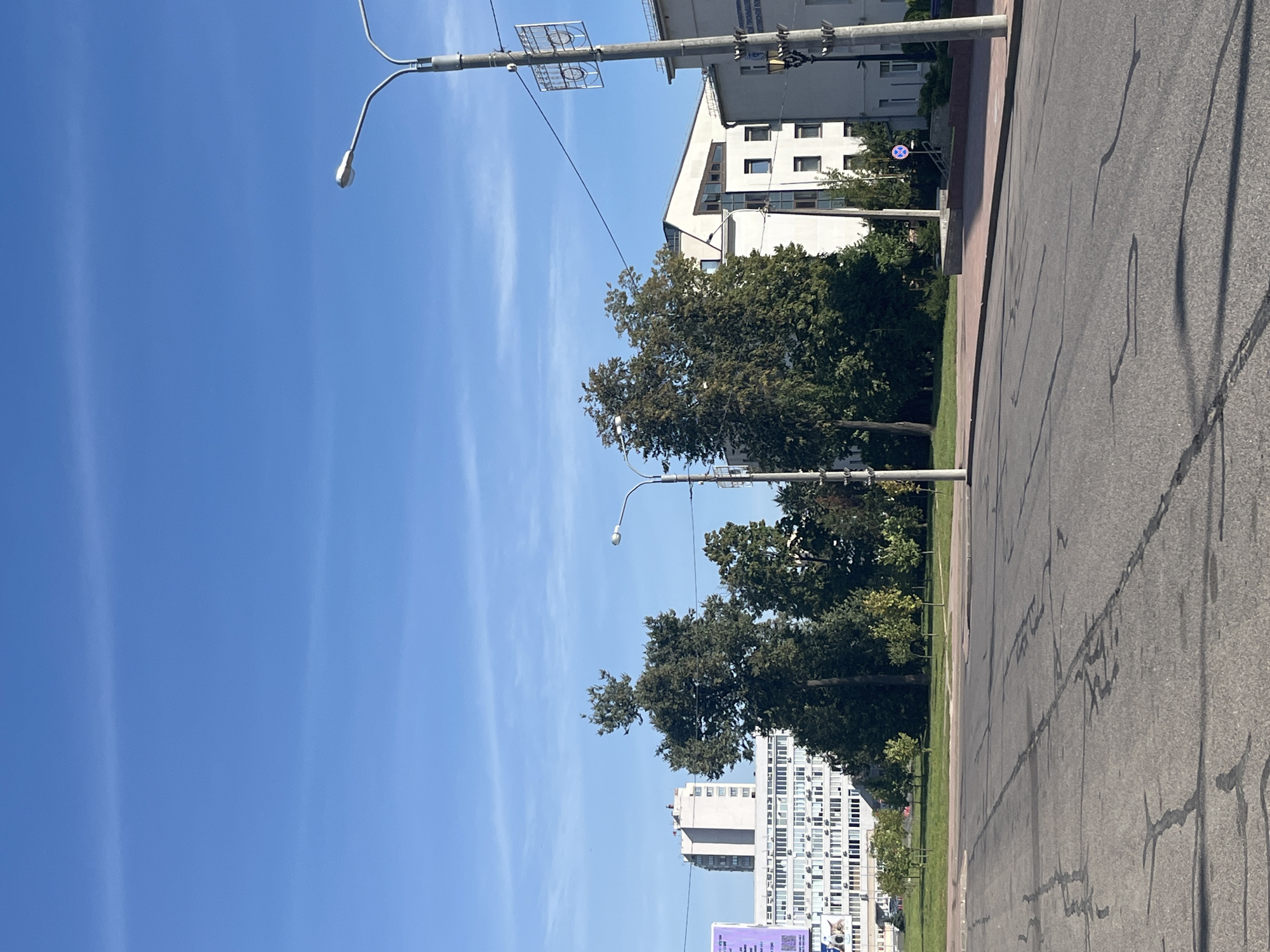 The image depicts a street scene in Minsk, Belarus, on a clear, sunny day. In the foreground, a paved road shows signs of wear with numerous cracks and patches. A sidewalk with a red brick border runs along the right side of the road. Beyond the sidewalk, a grassy area is visible, dotted with young trees and larger, mature trees.
Dominating the background is a large, multi-story white building with many windows, possibly an administrative or educational institution. To its left, another taller, white building with a distinctive tiered structure is visible. Several streetlights are present, with one prominent lamppost on the right side of the image featuring a decorative metal frame near its top. The sky is a bright, cloudless blue. No people or vehicles are present in the frame. The overall impression is of a quiet urban setting.
