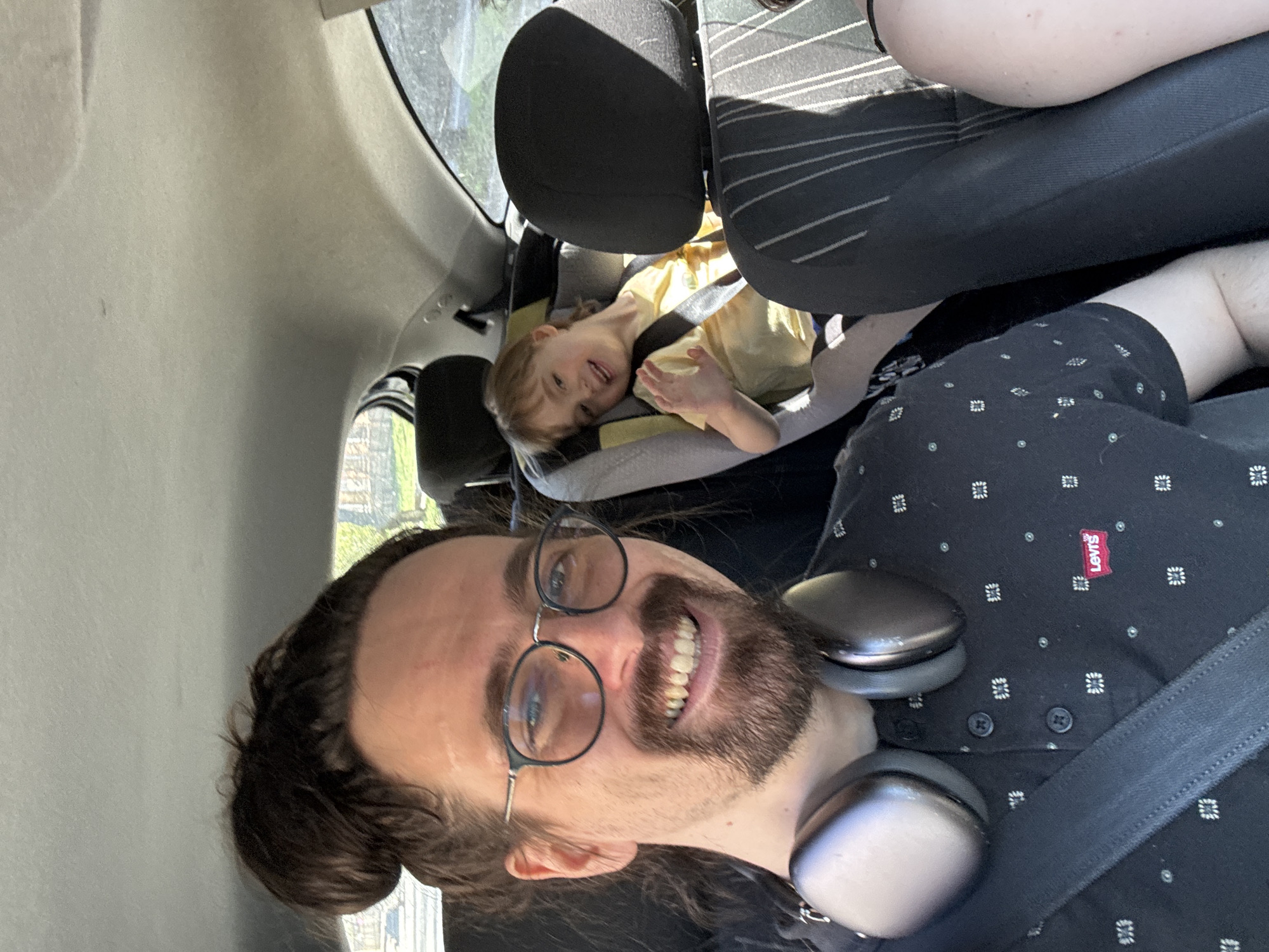 Inside a vehicle, a man with a beard and glasses, wearing headphones and a patterned shirt, smiles towards the camera. He is wearing a seatbelt. Behind him, a child in a yellow shirt sits secured in a car seat. The interior of the car is visible, including the seats and window. The location cannot be determined as Riga, Latvia, based on visual information.
