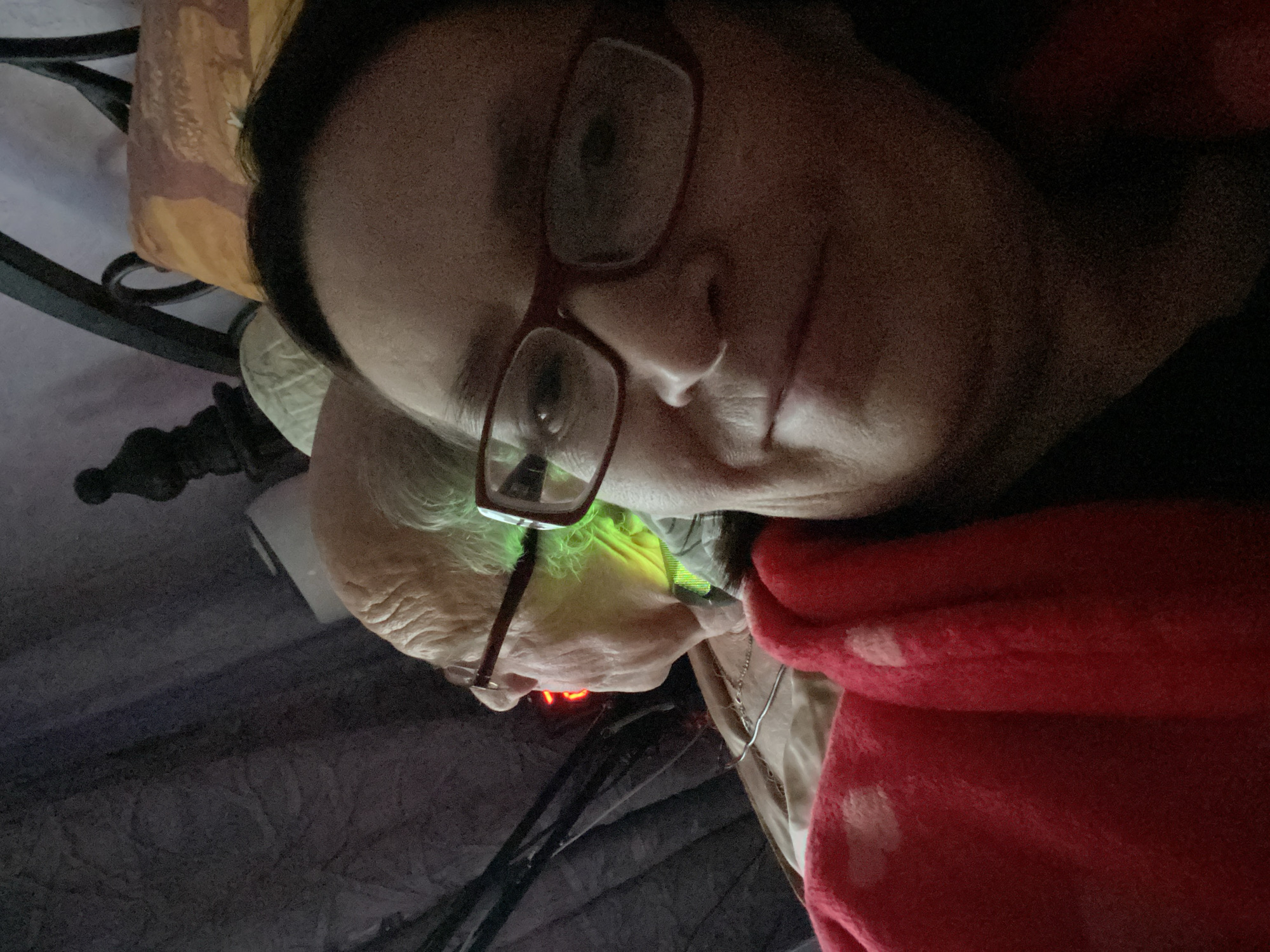 The image shows two individuals lying in what appears to be a bed. The person in the foreground has dark hair and glasses with red frames. A red blanket is draped partially over them. The second person, situated in the background, is elderly with wrinkled skin and also wears glasses. There's a green light source illuminating their face, and an orange/red light is visible near their neck. The setting includes a decorative pillow with a patterned cover, and part of a metal bed frame. There is no information in the image to support the location of Novokuznetsk, Russia.