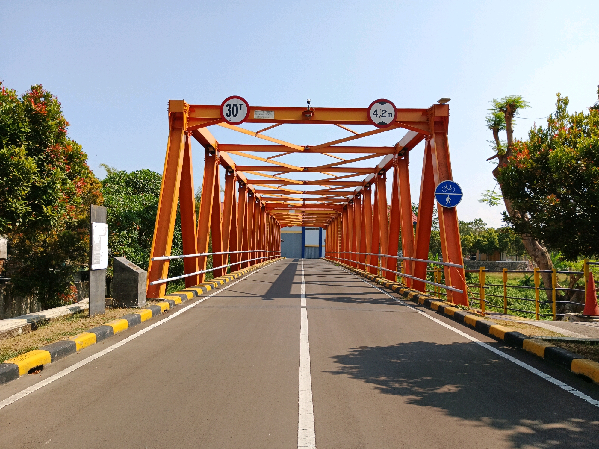 The image shows an orange truss bridge with a road traversing it. The bridge is illuminated by bright sunlight, casting shadows on the asphalt road. On the bridge, there are traffic signs indicating a weight limit of 30 tons and a height limit of 4.2 meters. There are also signs for bicycles and pedestrians. The bridge is flanked by greenery, including trees with lush foliage, and a clear blue sky overhead. In the background, a building with a blue and white facade is visible. The road is marked with white lines, dividing it into lanes. Yellow and black striped barriers line the sides of the road. The scene appears to be a typical urban landscape in Bandung City, Indonesia, possibly a scenic route or a passage over a river or a lower road.