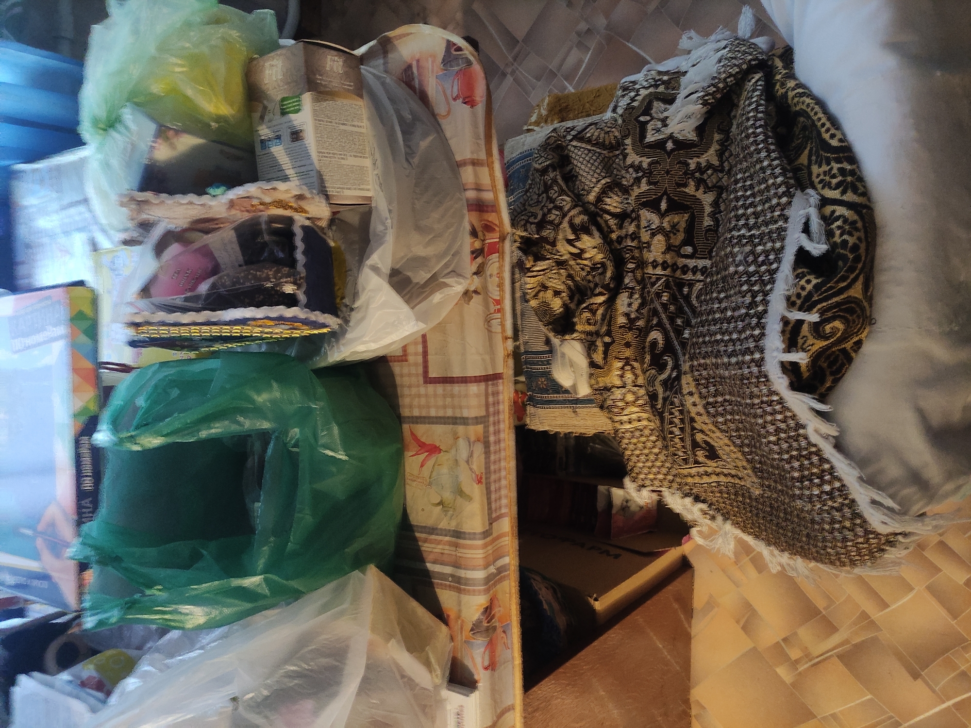 The image depicts a cluttered indoor scene, with various objects piled on what appears to be a low table or shelf. There are plastic bags, one green and one transparent, containing indistinct contents. A cardboard box with the word "Fit" is visible. A decorated object, possibly a cake or a similarly adorned item, is present. A blanket with a patterned design and fringe is draped over a white pillow. A table or shelf with a floral design is also visible. Given the prompt's request for the location, the image is likely taken in Pavlodar, Kazakhstan, although there are no distinctive features in the image to confirm this.