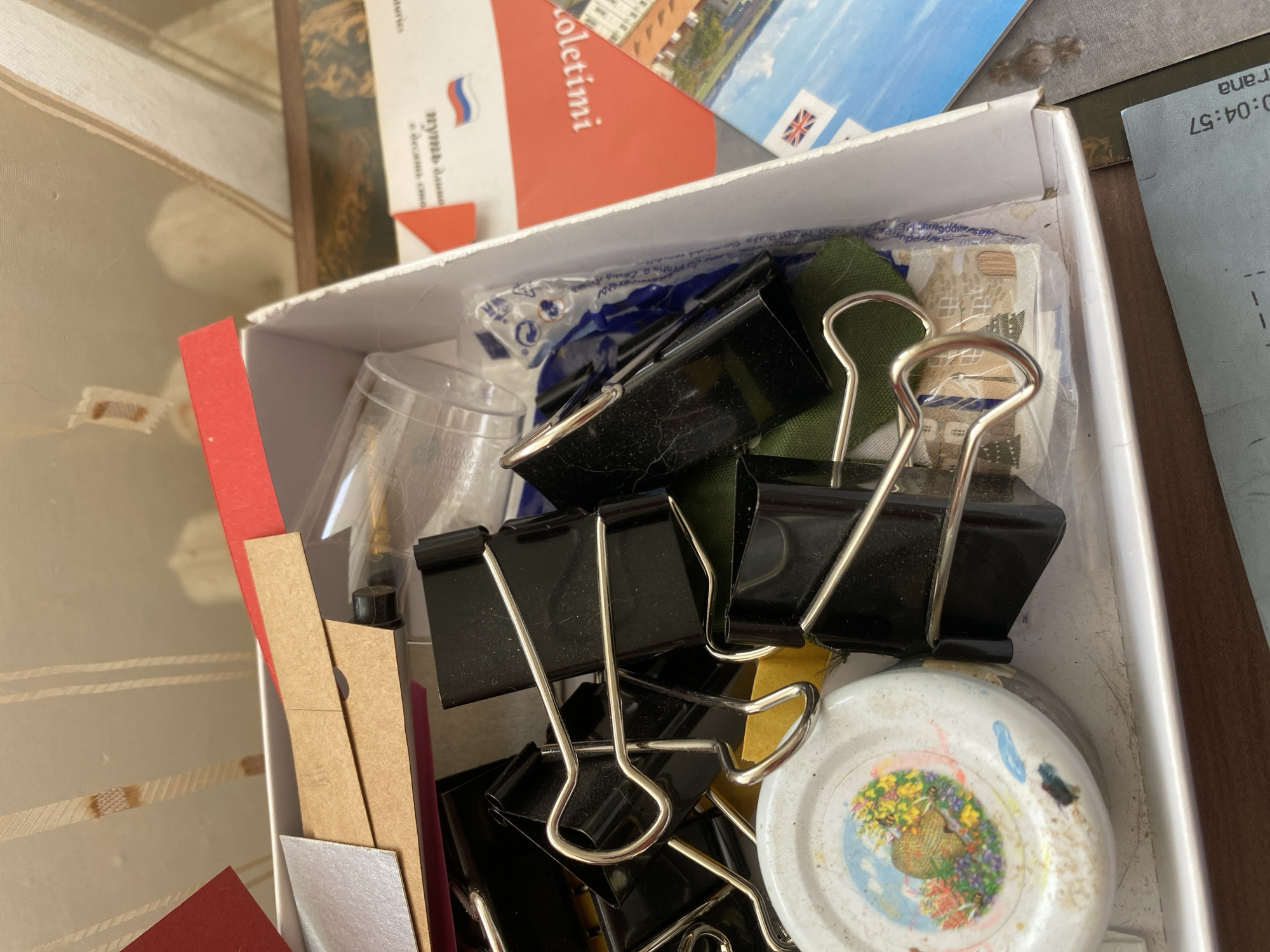 The image shows a close-up, high-angle view of an open white cardboard box filled with various craft supplies and office stationery. Dominating the frame are several large black binder clips with silver metal handles. Among these are also some pieces of colored paper, including red and tan, and a sheet of metallic silver paper. A small, round white object with a colorful illustration of a bee and flowers is partially visible on the left side, possibly a lid or a decorative element. Further in the box, a clear plastic cup and some plastic packaging are visible. In the background, a portion of a postcard or pamphlet featuring a city skyline and flags, including the Union Jack, is present. Another document with Cyrillic text and a stylized flag is also visible. The contents appear to be stored loosely within the box. The overall environment suggests a personal crafting or office space. The location of Bílina, Czechia is not depicted in the image.
