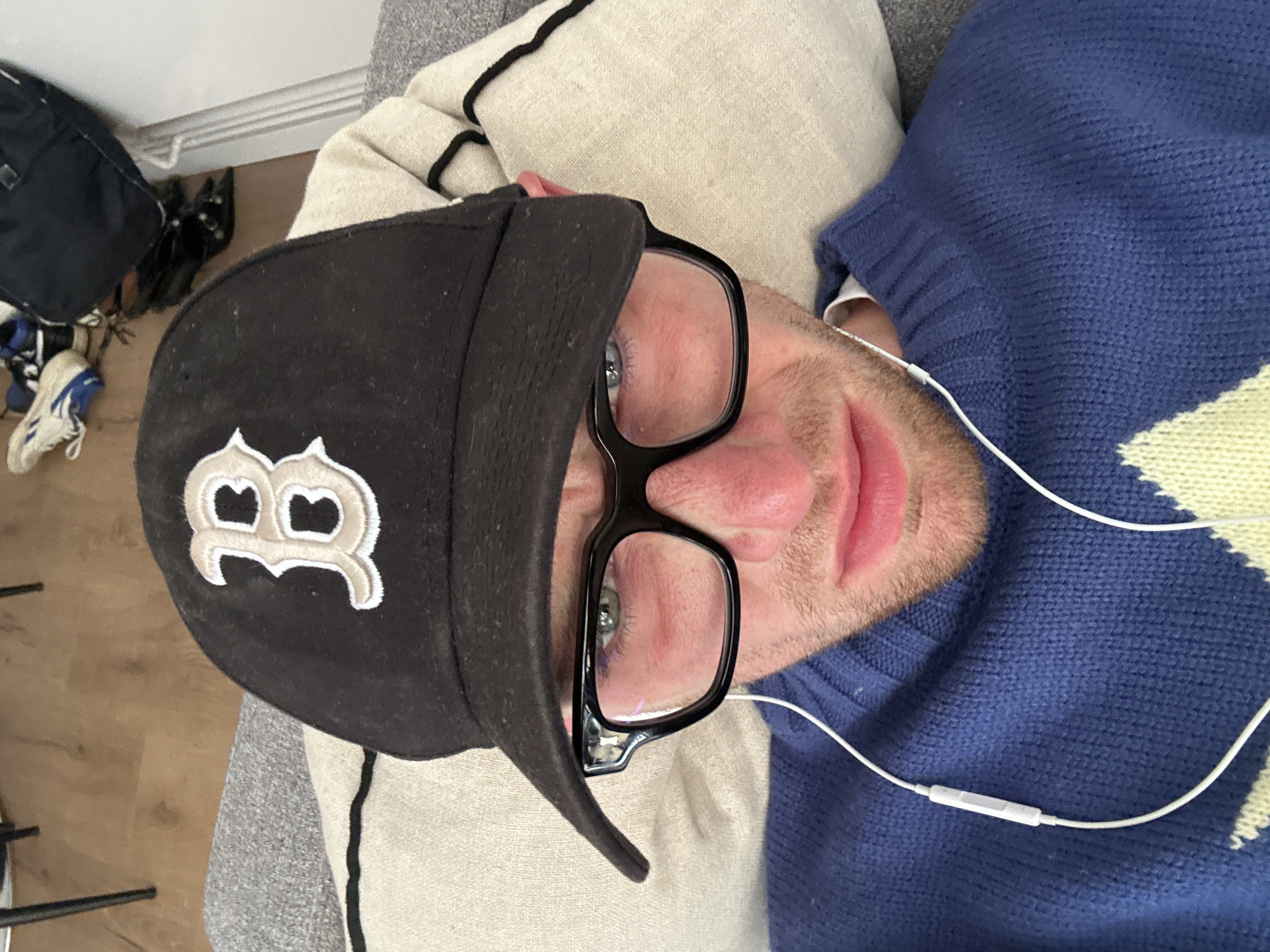 A man is lying down, possibly on a couch, wearing a black baseball cap with a white "B" logo. He is wearing eyeglasses with black frames and has white earbud cords extending near his face. The man has fair skin and short facial hair, and is wearing a blue sweater with a yellow diamond pattern. In the background, there is a wooden floor and a collection of items including a black backpack, shoes, and what appears to be a beige pillow. The location cannot be determined as being London, United Kingdom based on the provided images.