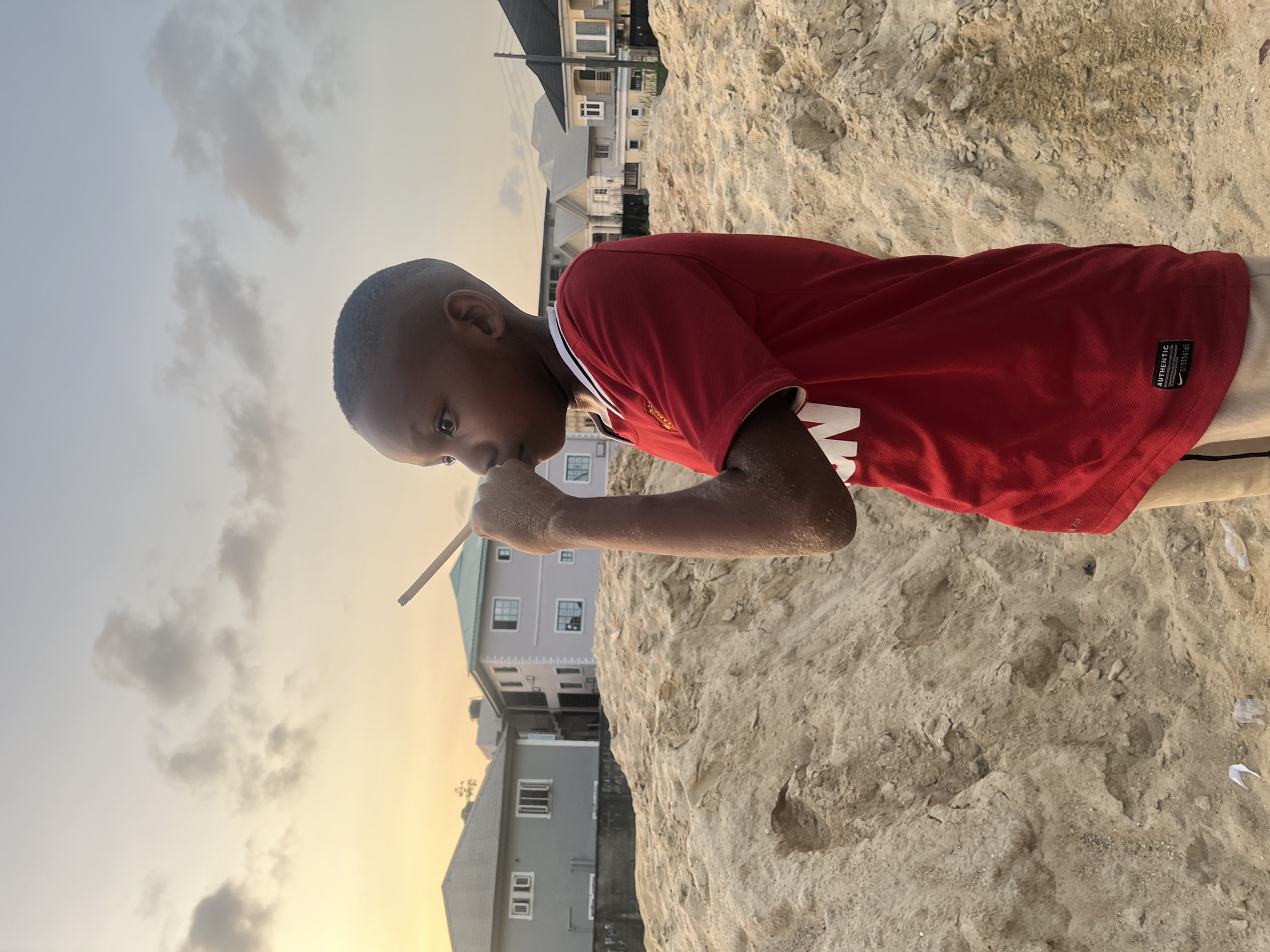 An outdoor scene captured at dusk or sunset in Iranla, Nigeria, depicts a young child of dark complexion, approximately 5-8 years old, standing in profile facing left. The child wears a red short-sleeved t-shirt with white trim and light-colored shorts. Their right arm and hand are visibly coated in light brown sand. The child's right hand is raised towards their mouth/nose area, holding a slender light-colored wooden stick between the thumb and index finger, extending upwards. The child's gaze is directed forward.
The foreground and midground are dominated by a large, irregularly shaped mound of light brown sand with visible indentations and disturbed areas. In the background, beyond the sand pile, several modern residential buildings are present under a sky transitioning from light blue to warm orange and yellow, with scattered grey clouds. Buildings include a grey two-story house with a dark roof on the left, and a purple/grey two-story house with a teal roof and external staircase in the center-right. Additional residential structures with varying roof colors are visible further right.