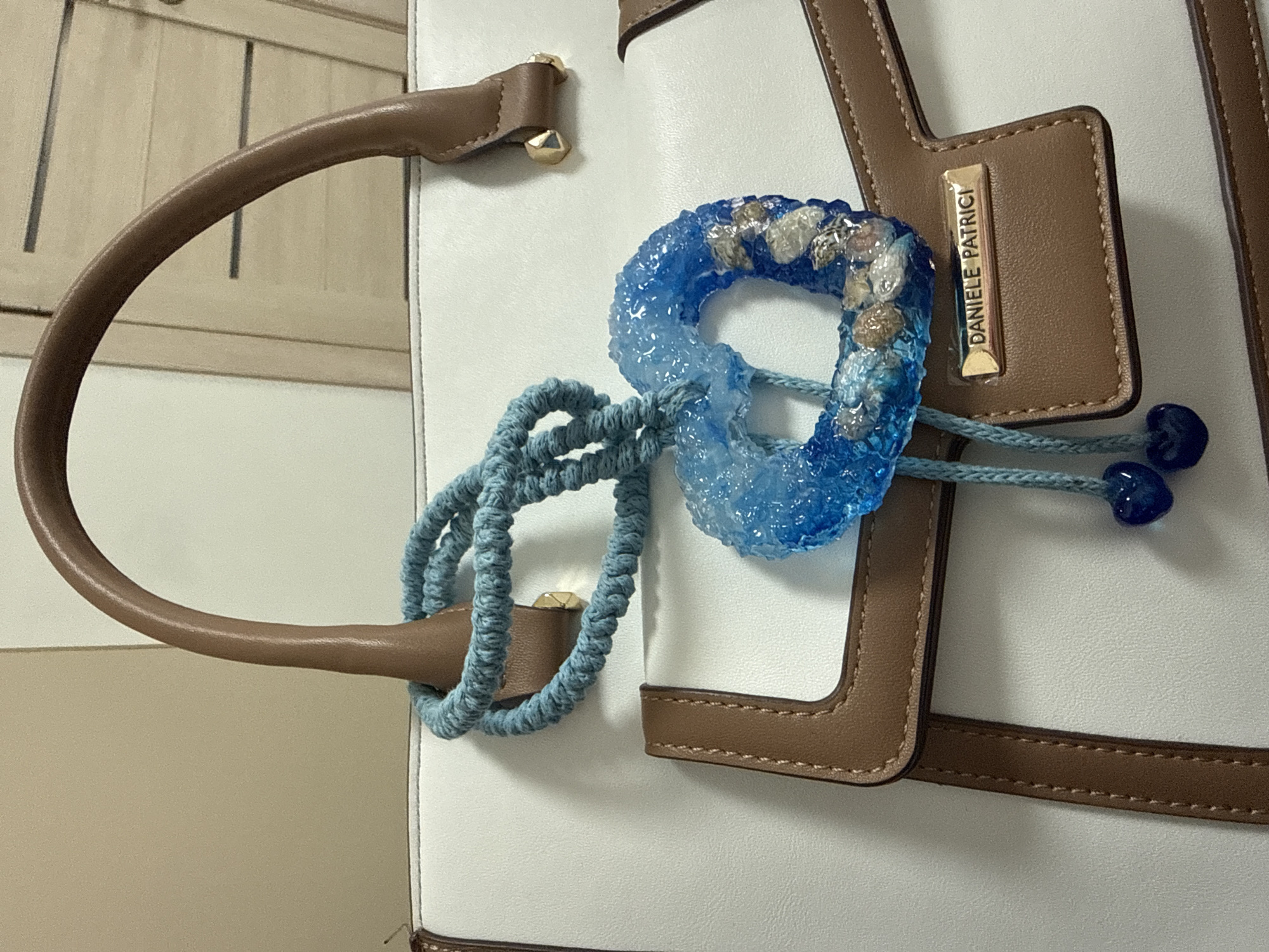 The image shows a close-up of a white leather tote bag with brown trim. A blue braided cord is looped through the handle, and a blue resin heart charm with small shells and white pebbles embedded in it hangs from the cord. Two small blue heart-shaped charms are attached to a blue string dangling below the heart charm. A rectangular gold-colored metal tag with the name "DANIELLE PATRICI" embossed on it is affixed to the front of the bag. The background is a plain, light-colored wall. The image does not provide any information about the location being Vladivostok, Russia, or any specific context beyond the objects depicted.