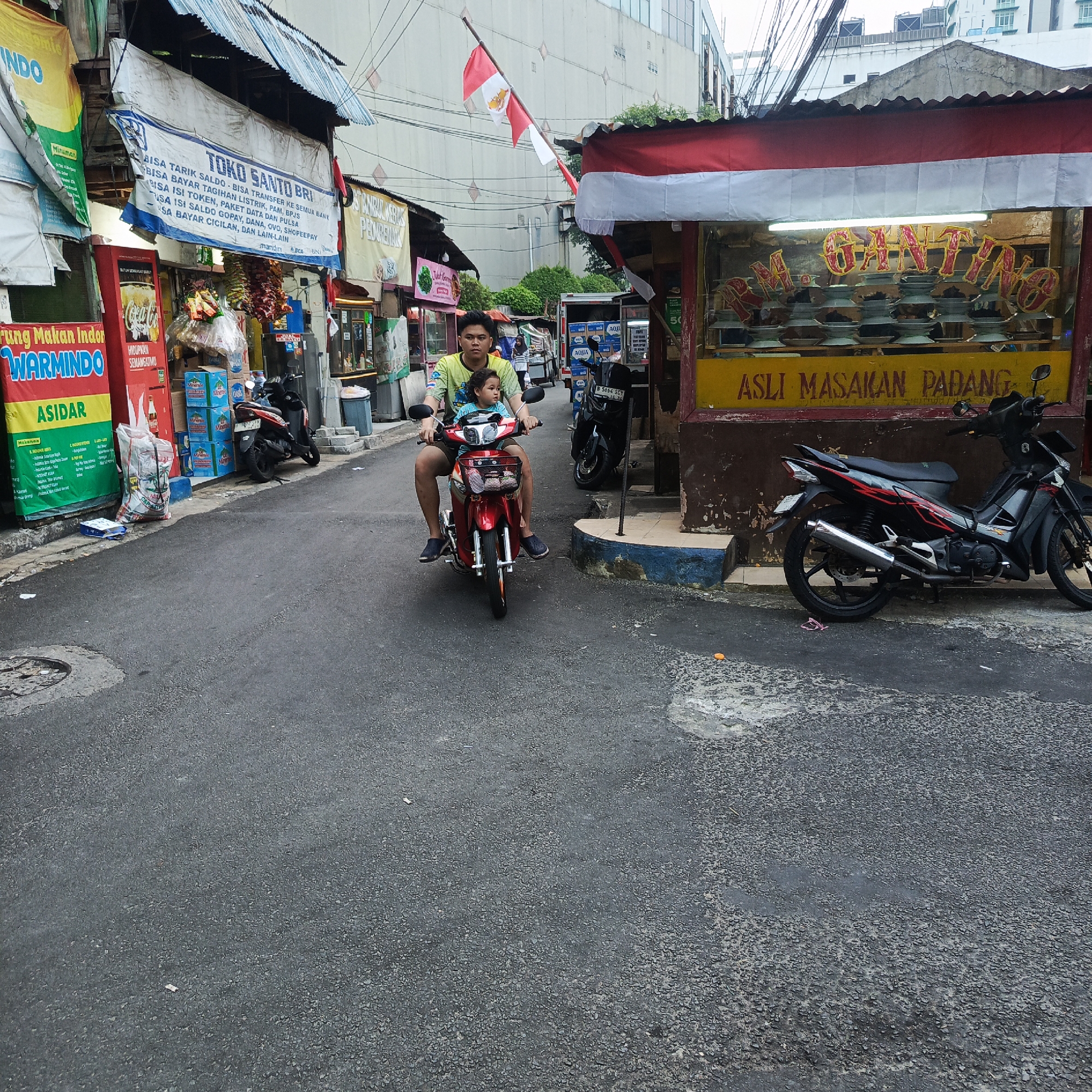 In South Jakarta, Indonesia, on a paved street lined with small businesses, a man and a young child ride a red motorcycle. The man, wearing a green and yellow patterned t-shirt and dark shorts, drives the motorcycle while the child, in a light blue shirt, sits in front of him. Both are looking forward. Behind them, other motorcycles are parked. To the right of the street, a food stall with a red and white awning displays food in bowls and has the words "ASLI MASAKAN PADANG" (Authentic Padang Cuisine) written in yellow on its facade. Indonesian flags are visible on poles above the street. Signs for "TOKO SANTO BRI" (Santo BRI Shop) and "WARMINDO" (Indonesian Food Stall) are also present. The environment appears to be an urban setting with buildings in the background.