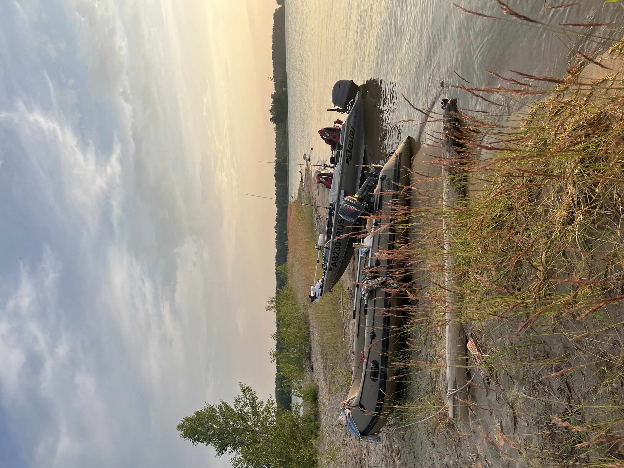 Two inflatable boats are moored on a sandy shore, with tall grasses in the foreground. The boat closest to the camera is a tan-colored inflatable with a shorter engine. Behind it, a second, larger boat, gray and white, is equipped with a trolling motor and appears to be carrying fishing rods. The water is calm, reflecting the soft light of a setting sun. In the distance, a treeline borders the horizon under a sky with scattered clouds. The scene evokes a peaceful, natural setting. The provided location, Borovoe, Russia, is a well-known resort area, and this image accurately depicts the characteristic landscape of its lake shores.