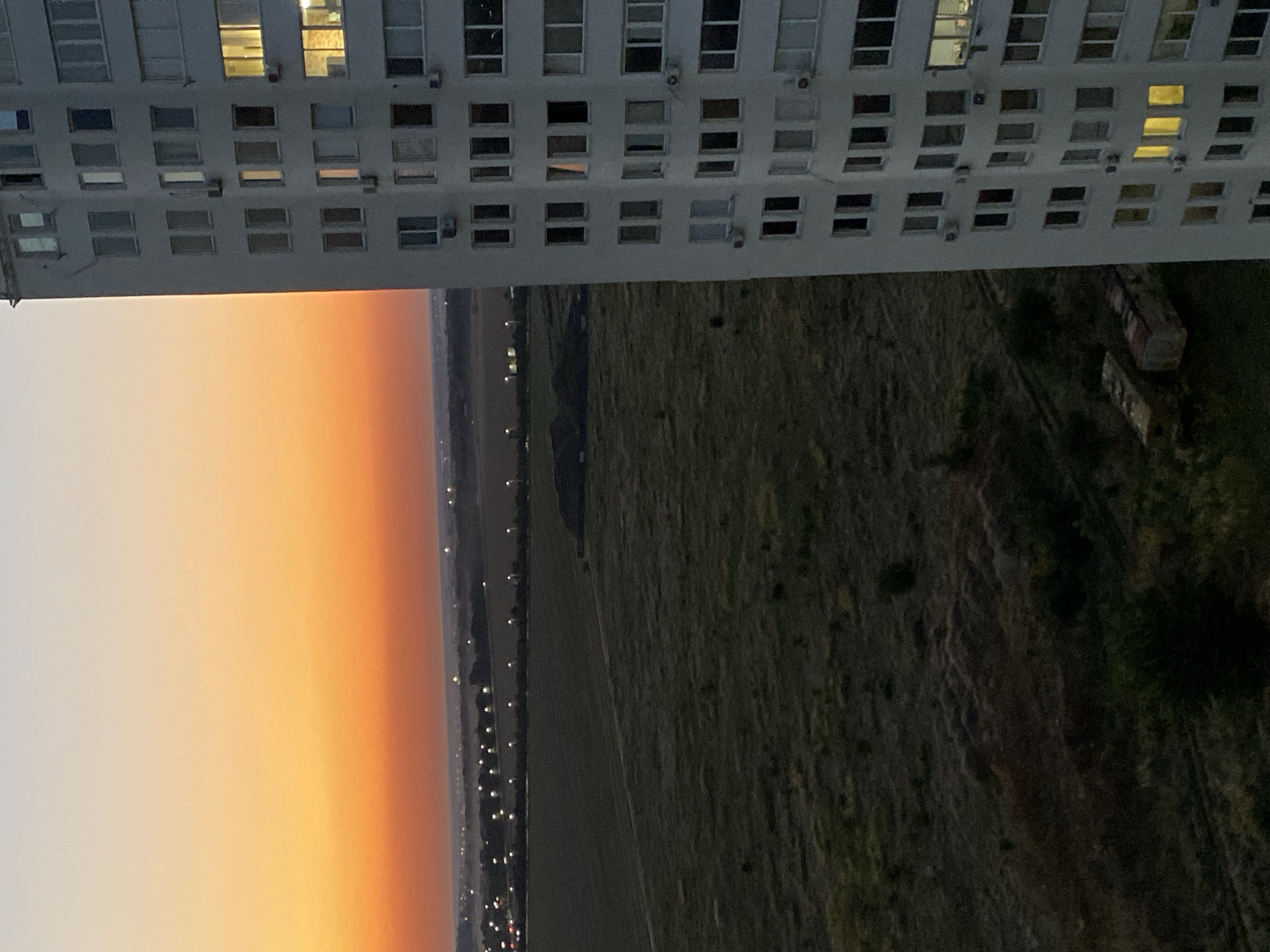 An exterior, vertical, medium shot captures a high-rise apartment building situated on the right side of the frame, angled towards the left. The building, predominantly white, features a curved facade and numerous windows, some of which are illuminated with a warm, yellow light. Several air conditioning units are visible mounted below the windows. In the foreground, the ground is covered in dark, dry vegetation, with two dilapidated, rectangular structures resembling old trailers or abandoned vehicles visible in the lower left quadrant. Beyond the foreground, a vast, open field extends into the mid-ground, transitioning to a distant landscape dotted with streetlights arranged in rows, indicating a settlement or town. The background is dominated by a vibrant sunset, with a gradient of colors ranging from a pale yellow at the top to a deep orange and red closer to the horizon. The overall scene suggests a view from a window or balcony overlooking a suburban or semi-rural area on the outskirts of Chornomorsk, Ukraine, during twilight. There are no people or animals visible in the image.