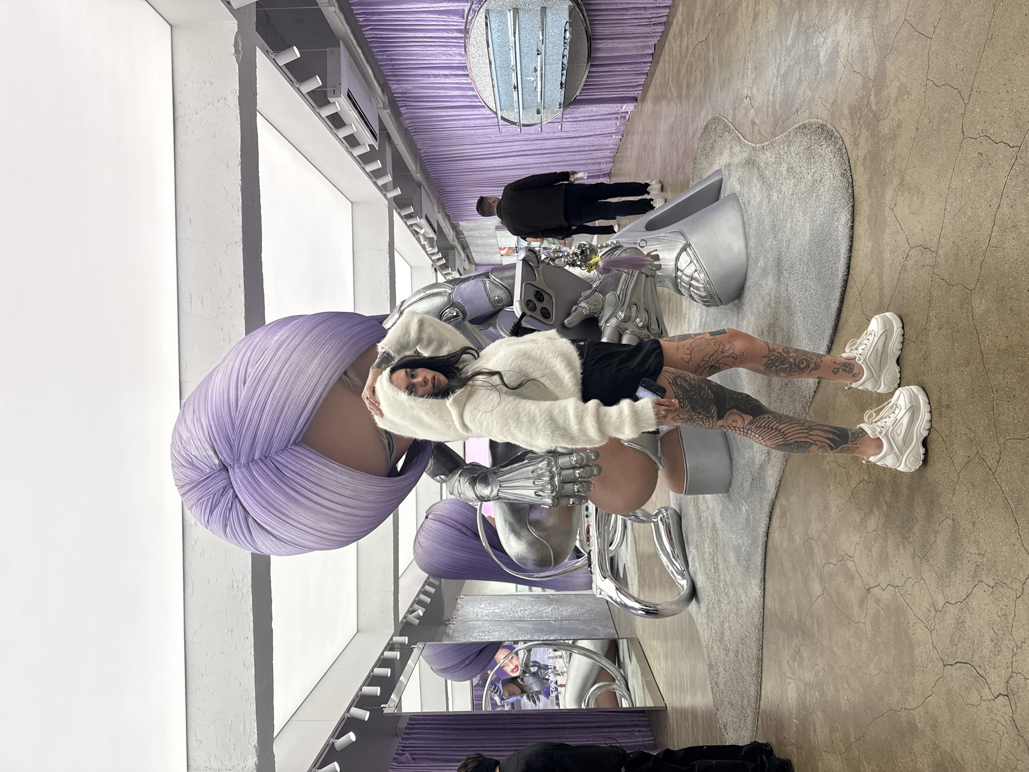 The image shows a person with a long-sleeved white furry coat, black shorts, and white sneakers. The person has tattoos on their legs. The person is interacting with a metallic silver chair structure that has a large lavender fabric adornment resembling a head wrap attached to it. The scene appears to be an installation or art exhibit. The background features white walls, a ceiling with lighting fixtures, and lavender-colored curtains. The floor has a gray rug and a textured beige surface. In the distance, other people can be seen observing the scene. Based on the visual details and style, the location might be an art or design space in Seoul, South Korea. The presence of the unusual chair structure and the stylized environment suggests that the setting is meant to be artistic and avant-garde.