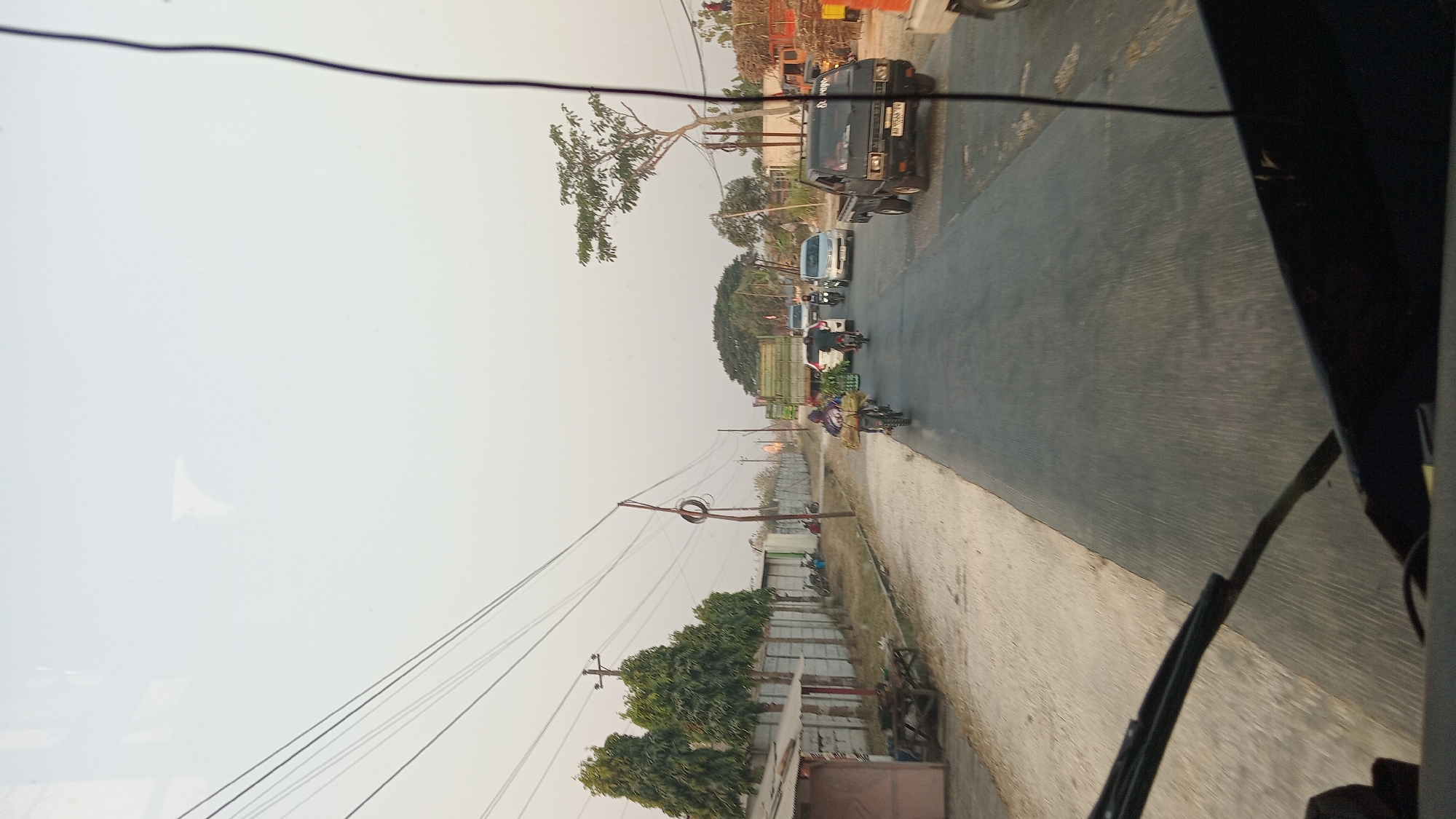 This is a photograph taken from inside a vehicle, looking out at a road scene in Klokah, Indonesia. The sky is overcast with a pale, hazy light, suggesting it is either early morning or late afternoon. The road is paved and appears to be a two-lane street with traffic.

On the left side of the road, there is a row of tall, slender trees in front of a grey concrete wall. Adjacent to the wall is a small, weathered building with a reddish-pink facade and a corrugated metal roof. A wooden table and chairs are positioned in front of this building. Further down the road on the left, there are several parked motorcycles and a person sitting near them.

The road itself is populated with various vehicles. A person on a motorcycle, carrying a load of green produce, is in the foreground on the left lane. Behind them, a white car is also moving. In the right lane, a black pickup truck with a license plate visible (partially obscured but reads "AD 8554 VV") is a prominent feature. Further ahead, a light-colored car and another motorcycle are visible. To the far right, a large truck loaded with what appears to be hay or dry vegetation is parked. In the background, more trees and structures are visible.

The image captures a moment of everyday life on a road in a rural or semi-urban area of Klokah, Indonesia, with a mix of local transportation and commercial activities. The presence of overhead power lines is also a notable environmental feature.