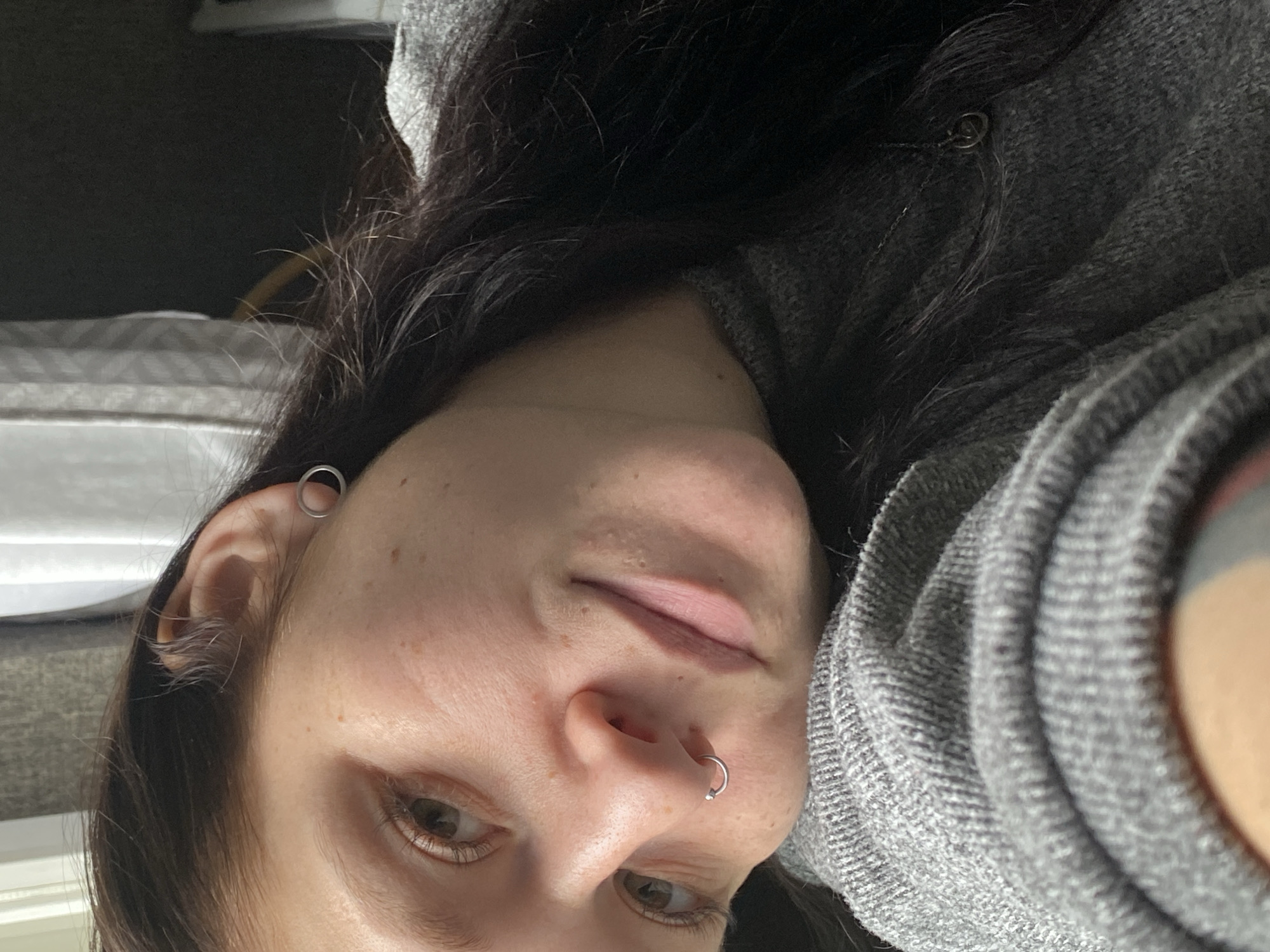 A close-up, slightly rotated, selfie of a person with fair skin, brown hair, and brown eyes. The person is wearing a gray, textured garment, possibly a sweater or hoodie. The person has two visible silver piercings: one in the upper ear cartilage and one in the left nostril. The background is nondescript. No location information can be inferred from the image.