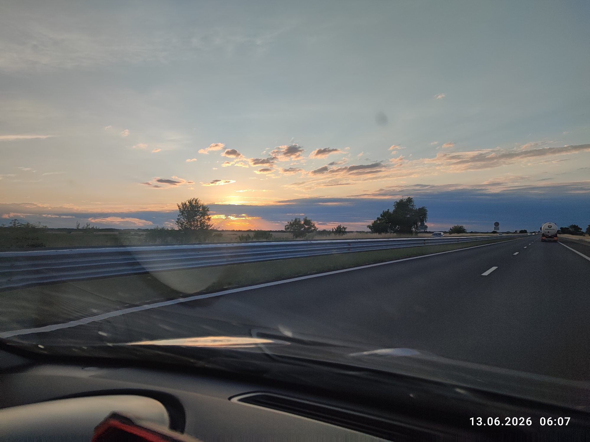 The image depicts a sunset scene from inside a vehicle on a highway, with the timestamp "13.06.2026 06:07" visible in the lower right corner. The highway stretches into the distance under a sky filled with clouds illuminated by the setting sun. The foreground shows the dashboard and windshield of the vehicle, reflecting the environment. To the left of the highway, a metal barrier separates the road from a grassy area with trees and vegetation. In the distance, a tanker truck is visible on the highway, and a signpost stands on the right side of the road. The location is stated to be Malo Konare, Bulgaria, suggesting this highway is situated near that area.