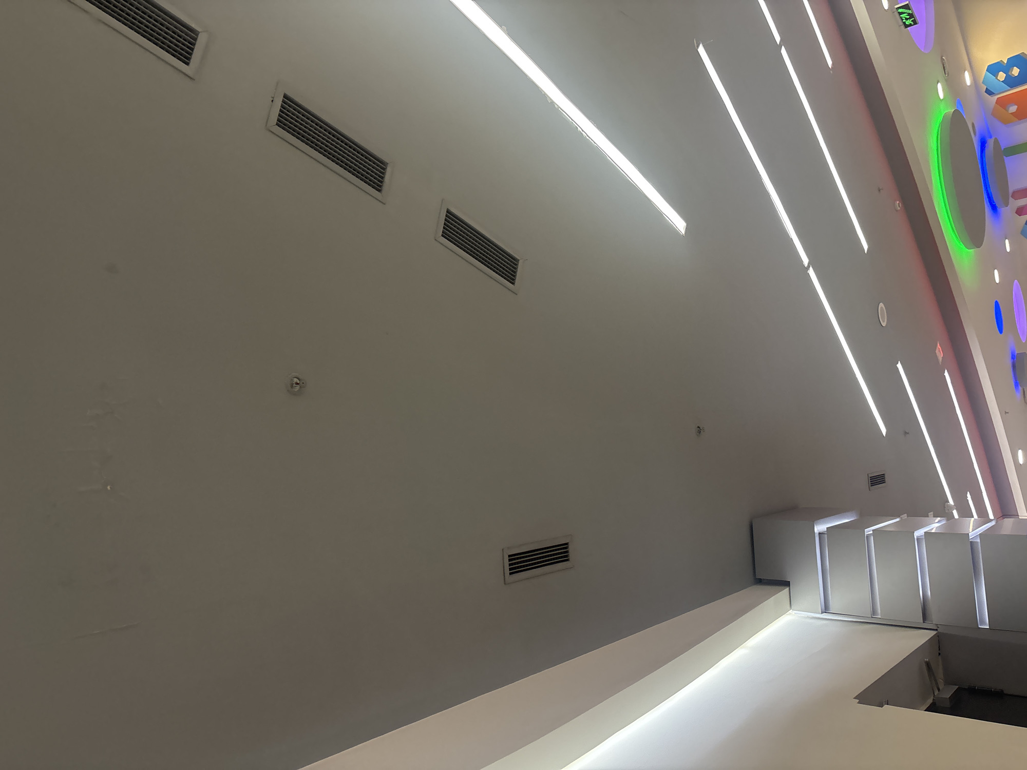 This media file captures an interior architectural space characterized by a bright, modern aesthetic. The scene primarily features a light gray ceiling and upper wall, illuminated by multiple long, linear LED light fixtures arranged diagonally across the ceiling. Several rectangular air vents (HVAC diffusers) are visible on the ceiling and upper walls, along with small circular smoke detectors or sprinklers.
On the right side of the image, the space incorporates striking geometric white structures that appear as steps or display platforms, with integrated lighting illuminating their edges. Adjacent to these, a wall features various brightly colored, backlit decorative shapes, including circles, rectangles, and abstract forms, glowing in green, blue, purple, and pink hues. Visible text on one of the green-lit shapes at the top right corner appears to be "WIFI" with a signal icon.
The setting suggests a contemporary commercial building, possibly a shopping mall, entertainment complex, or a public area designed for visual engagement. No people are present. The strong artificial lighting indicates an indoor environment, making the time of day and weather irrelevant. Subtle stains or discoloration can be observed on the upper left wall. The overall impression is one of a clean, modern, and visually dynamic environment.