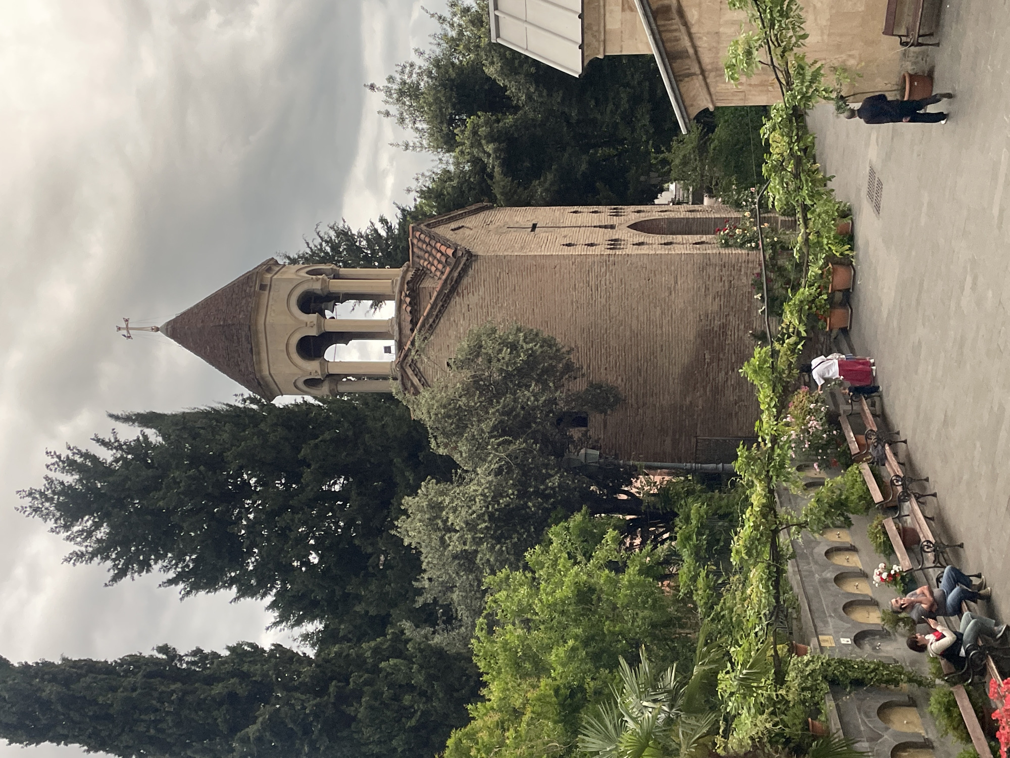 The image shows a bell tower in Tbilisi, Georgia, surrounded by trees and greenery. The tower is made of light brown brick and has a copper-colored roof with a cross at its peak. The tower's top section features arched openings. A paved walkway runs along the side of the tower, with benches and flower pots lining a low wall. People are seen walking and sitting on the benches. The sky is overcast.
