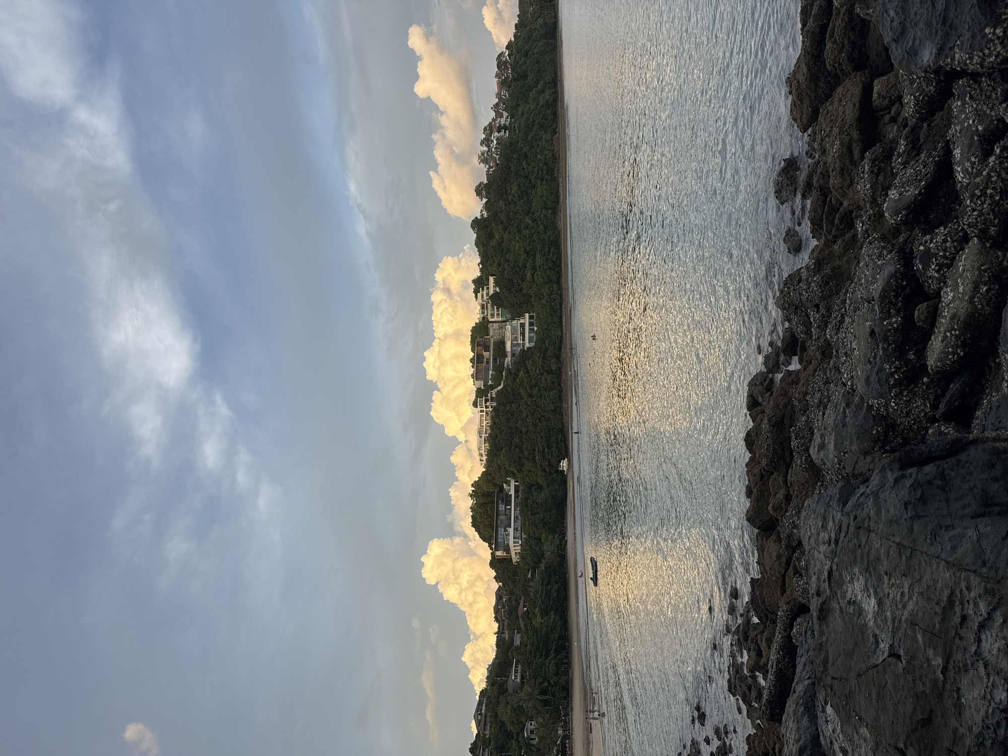 The image depicts a tranquil beach scene at dusk, likely located on the coast of Wichit, Thailand. The foreground is dominated by a cluster of dark, weathered rocks, some encrusted with barnacles, leading into the gently lapping water. The sea, reflecting the soft hues of the setting sun and sky, stretches towards a verdant hillside. Perched on the hillside are several modern, white houses with clean architectural lines, nestled amidst dense green trees. The beach itself is visible in the midground, with a few scattered individuals walking along the shore and wading in the shallow water. A small boat rests on the sand, and a single bird stands near the waterline. The sky above is a blend of soft blues and greys, punctuated by dramatic, luminous clouds that appear to glow with the last rays of sunlight. The overall impression is one of serene, affluent coastal living at the end of the day.