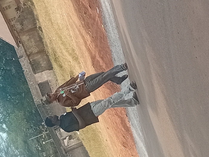 The image displays two males walking on a paved road in Ayangba, Nigeria. The male on the left, dressed in a black short-sleeved t-shirt and light grey baggy jeans, holds a small, light-colored rectangular object in his left hand and looks towards the male on the right. The male on the right wears a camouflage baseball cap, a brown long-sleeved shirt with a graphic design, and dark grey jeans. He holds a clear plastic bottle with a green cap in his right hand and a light blue rectangular object in his left hand, his mouth open, indicating speech. The foreground features a dark grey asphalt road, bordered by a strip of lighter gravel and a reddish-brown earth embankment with dry, yellowish-green grass. The background includes concrete block walls, some appearing unfinished, dense green foliage, and the corner of a building with a reddish-brown sloped roof.
