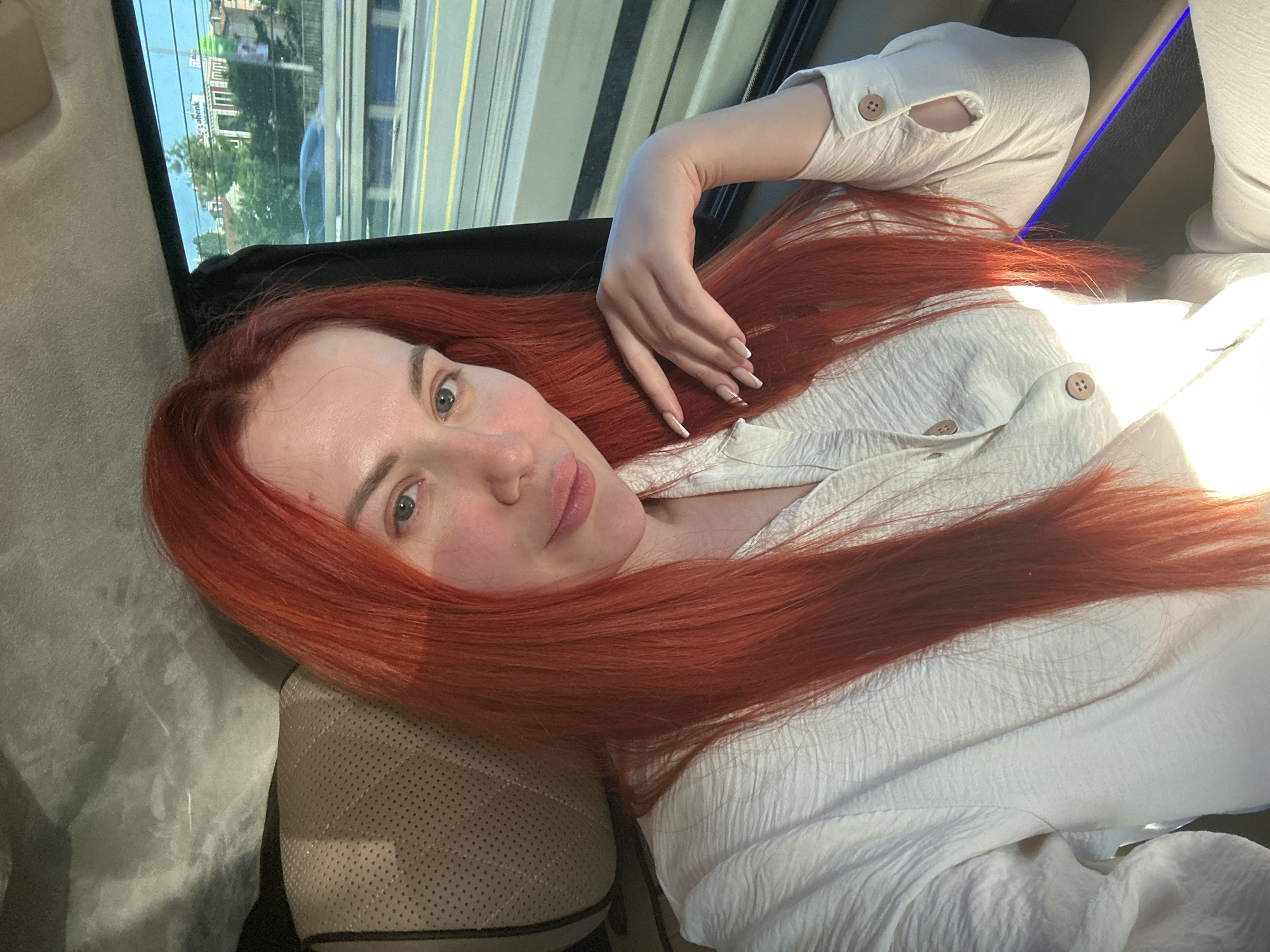 A woman with long, bright red hair is sitting in the back seat of a vehicle. She is looking at the camera and resting her chin on her left hand. She is wearing a white, textured shirt with buttons. The interior of the vehicle appears to be luxurious, with leather seats and ambient lighting. Through the window, a cityscape is visible, suggesting the vehicle is in motion. The scene is lit by natural light, casting shadows on the woman's face. The background outside the window shows buildings and trees, consistent with a city environment. The woman's nails are manicured and painted white.