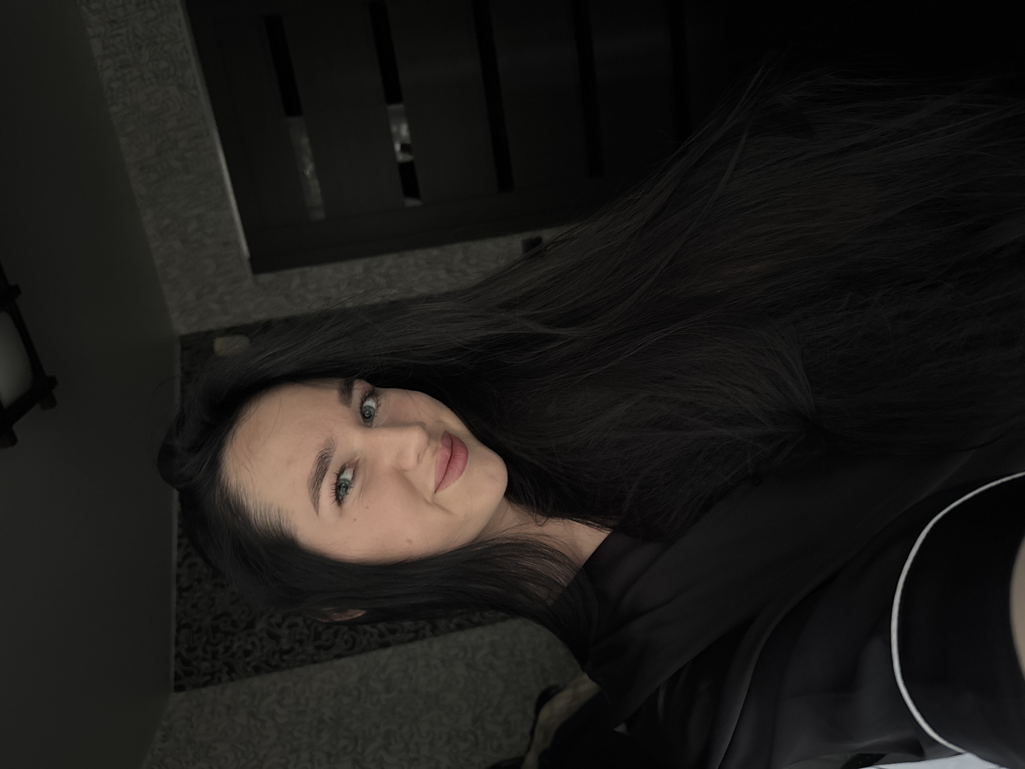 In a dimly lit room, likely an apartment in Novosibirsk, Russia, a young woman with long, dark hair and striking blue eyes is captured in a close-up, semi-profile shot. She is wearing a black robe with white piping. Her gaze is directed slightly off-camera, and she has a subtle, inviting smile. The background is largely obscured by shadow, but appears to include a textured wallpaper and a dark doorway or closet. The overall impression is one of intimate, candid moment.