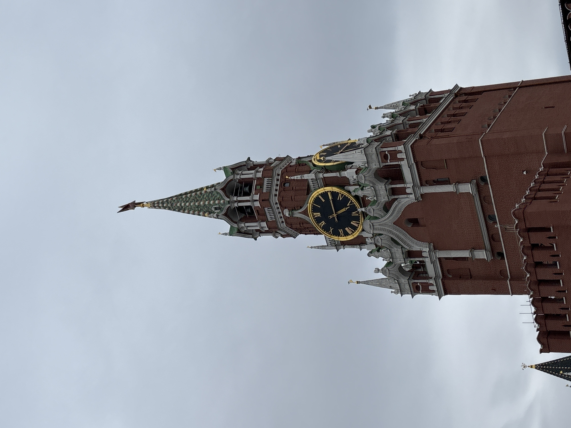 The image shows the Spasskaya Tower of the Moscow Kremlin in Moscow, Russia. The tower is red brick with white stone detailing. It has a large clock face with gold trim. The tower's pointed roof is green and decorated with a gold star. The sky is overcast and gray.