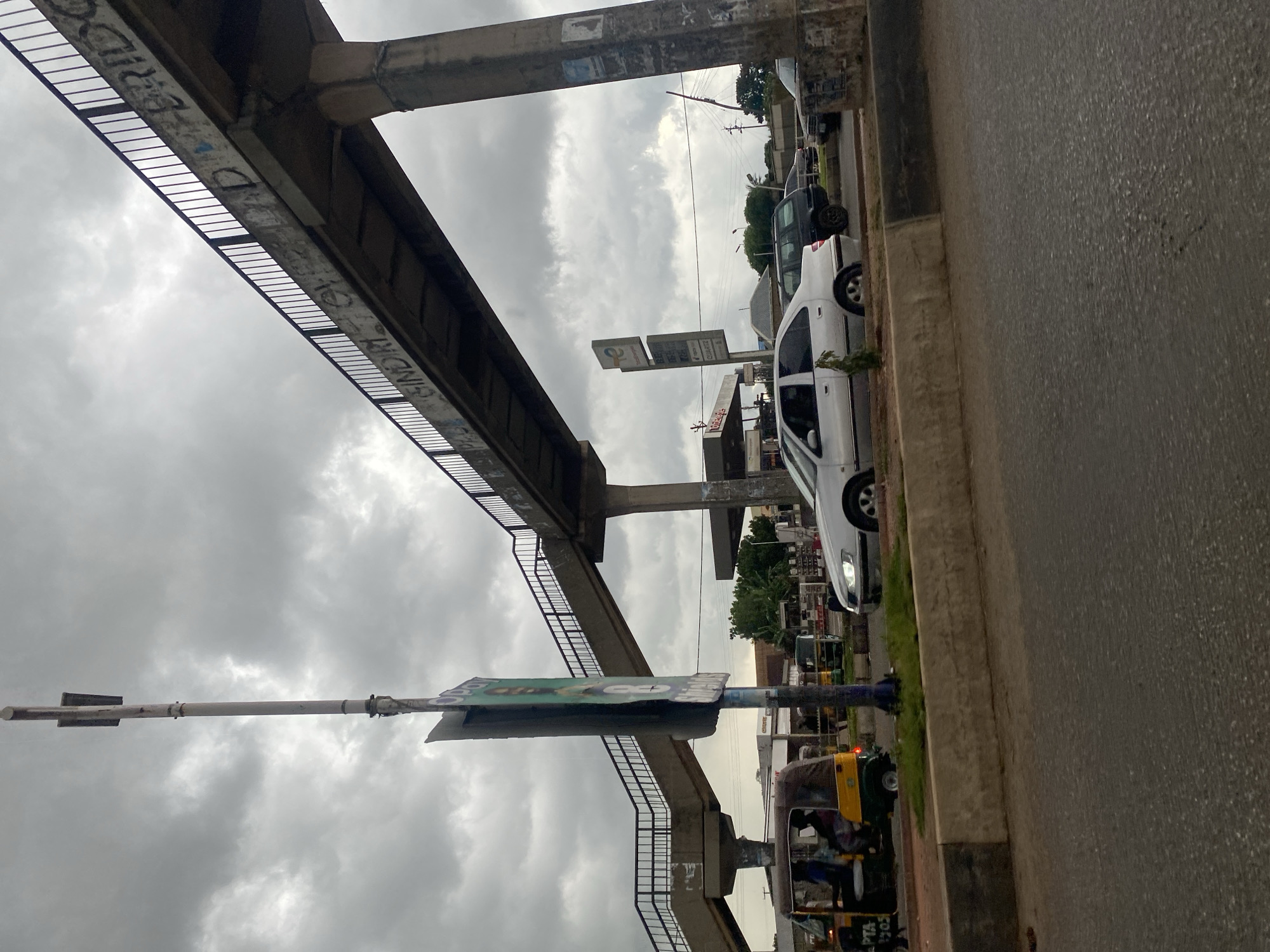 The image depicts an overcast day in Gura Top, Nigeria, featuring a concrete pedestrian bridge that spans over a street. The bridge's concrete surfaces are marked with graffiti. Below the bridge, a silver sedan is driving on the road, with other vehicles visible further down the street. A three-wheeled auto-rickshaw, also known as a Keke Napep, is in the left lane, carrying passengers. A tall, slender pole stands near the road, with a tattered green and white sign attached to it. In the background, a gas station with prominent signage and a canopy is visible, along with trees and power lines. The road surface in the foreground is asphalt, bordered by a concrete barrier.