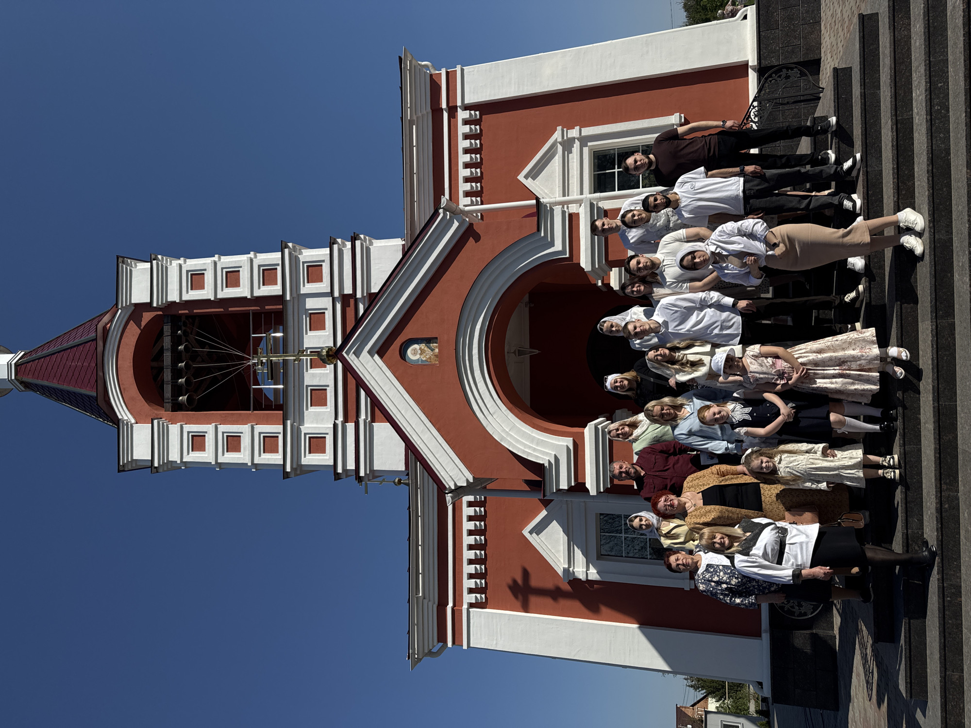 A group of approximately 20 people pose for a photograph on the steps of a red and white building. The building's architecture suggests it could be a church or religious structure, with a tall tower and arched entryway. The individuals in the photo vary in age and wear different styles of clothing. The sky is clear and blue.