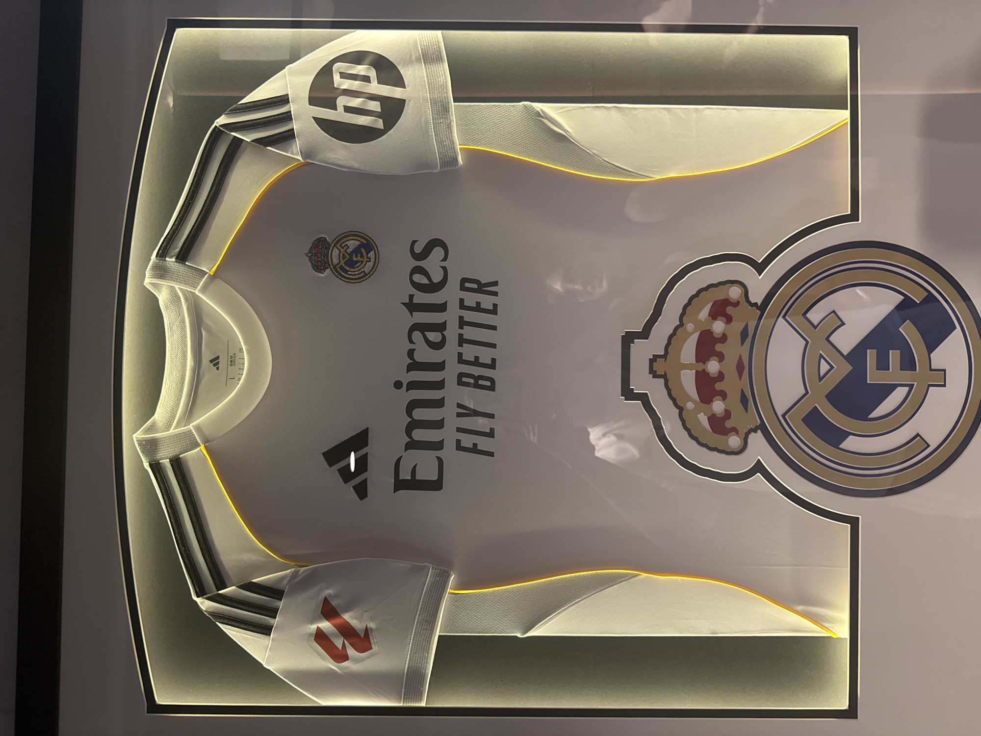 An Adidas Real Madrid football jersey is displayed in a frame with integrated lighting. The jersey is white with black Adidas stripes on the shoulders and sleeves. The Real Madrid crest is on the left chest, and the "Emirates FLY BETTER" sponsor logo is across the center. The HP logo is visible on the right sleeve. Below the jersey is the Real Madrid club crest in a larger format. The framing includes a light yellow glow around the jersey. There is no visible information indicating the location of this display in Kyiv, Ukraine.