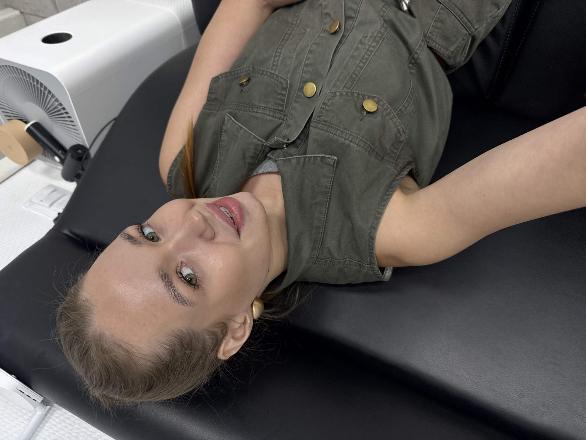 A woman is lying on a black surface, possibly a massage or medical table, in what appears to be a room with white walls. She is wearing a green sleeveless vest over a grey shirt and large, round gold earrings. Her hair is pulled back, and she is looking towards the camera with a slight smile. In the background, a white air purifier is visible. The image is taken from a high angle, looking down at the woman. There is no indication of the city of Chaikovsky, Russia, or any specific urban environment.