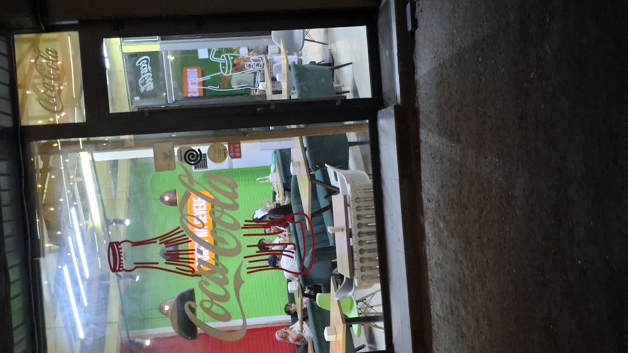 The image shows the exterior of a Coca-Cola themed cafe or restaurant in Shymkent, Kazakhstan, viewed through a glass storefront at night. The main display features a large, stylized red Coca-Cola bottle outline with the "Coca-Cola" logo in green script across its base. Inside, several patrons are seated at tables with green upholstery and light-colored tabletops. The interior walls are painted bright green, and string lights are visible in the upper ceiling area. Various Coca-Cola branding elements, including a smaller logo on a sign above the entrance and a graphic of a Coca-Cola glass with a straw, are present. The cafe appears to be open for business, with tables and seating arranged for customers. The lighting suggests it is evening or nighttime. The exterior pavement in front of the establishment is visible.