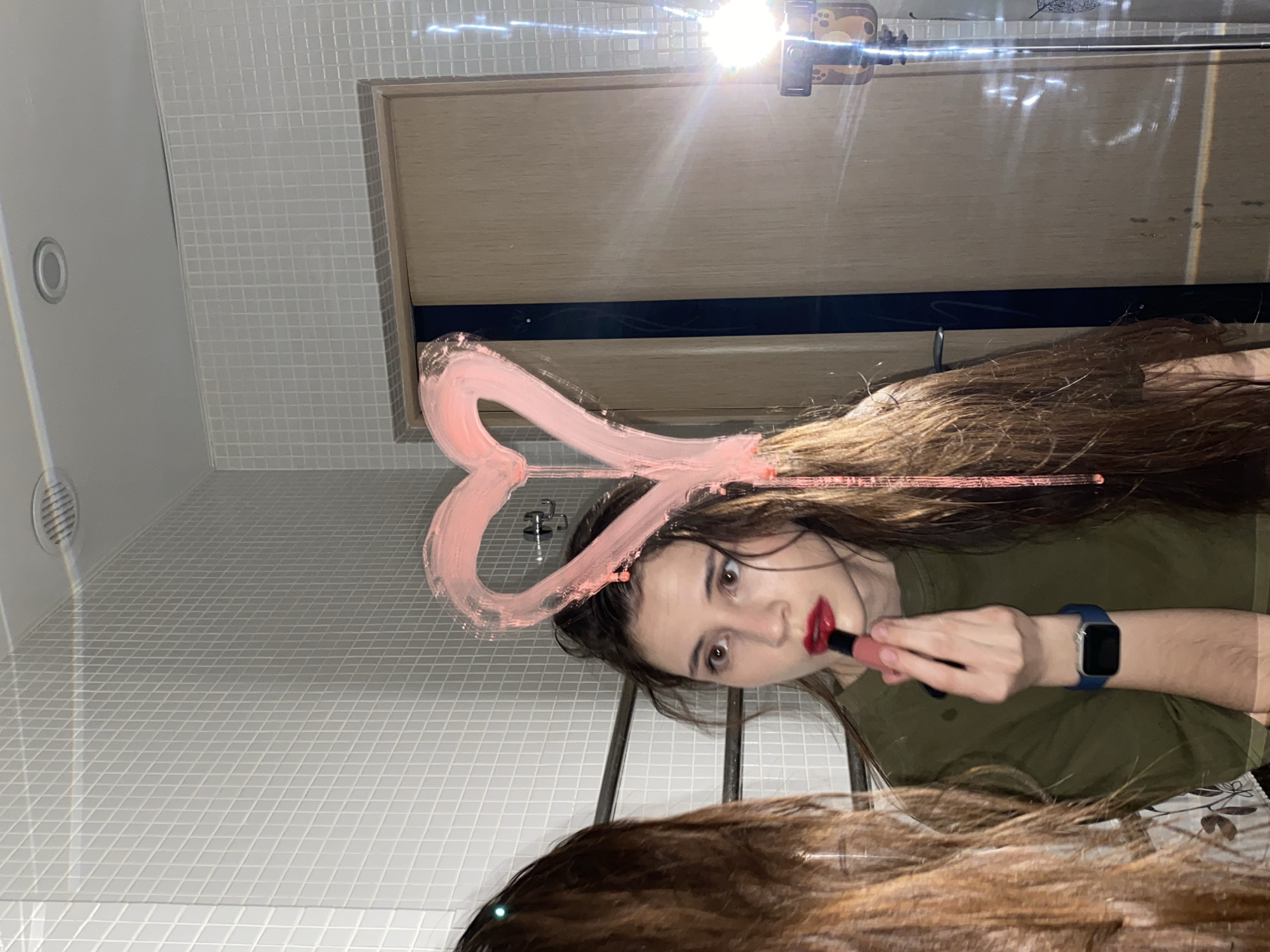 The image, taken in Votkinsk, Russia, presents a dynamic reflection of a young woman in a mirror, rotated 90 degrees clockwise. The woman, with long, dark brown hair, wears an olive-green shirt and a blue smartwatch on her left wrist. She is in the process of applying red lipstick, her gaze directed intently towards her reflection. A prominent pink heart, seemingly drawn with lipstick, frames the upper left side of her head on the mirror's surface.
The setting, viewed through the reflection, features small, white square tiled walls, indicative of an indoor space like a bathroom. Behind the woman, a wooden door or cabinet is reflected, above which a bright light fixture with a patterned bracket causes a significant lens flare across the upper portion of the mirror. Her abundant hair cascades downwards, partially obscuring the lower part of the reflection. The scene is well-lit, likely by artificial light, given the strong light source. No visible text is present.